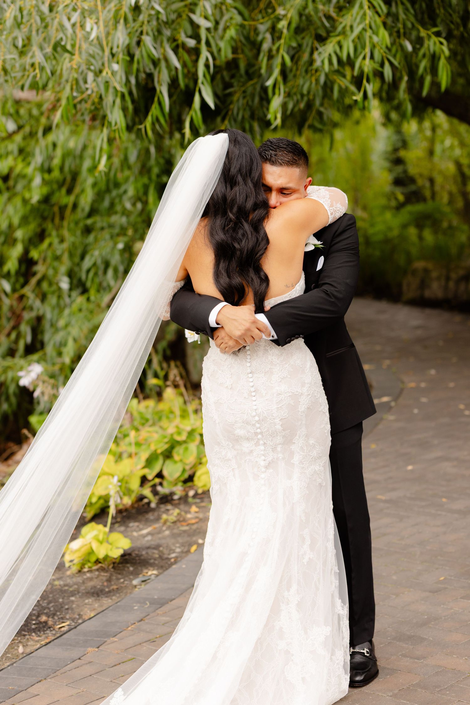 A newlywed couple shares an intimate embrace on a garden path with cascading willow branches in the background.