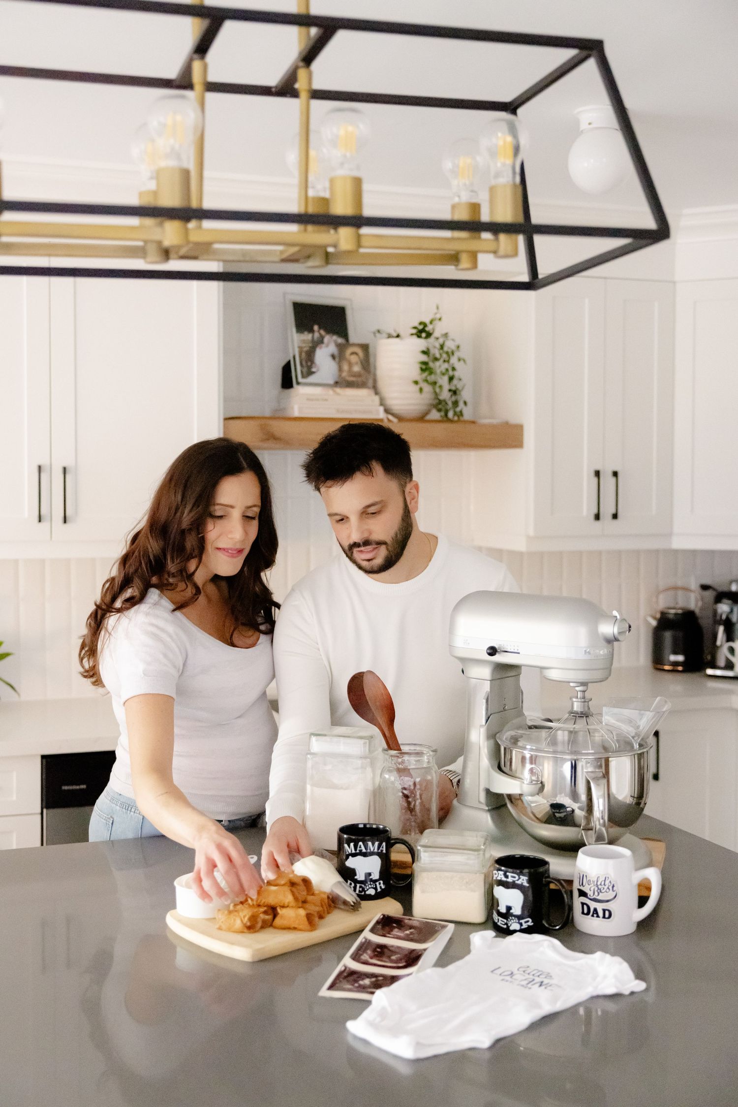 A couple prepares food together in a modern white kitchen with a stylish black and brass chandelier overhead.
