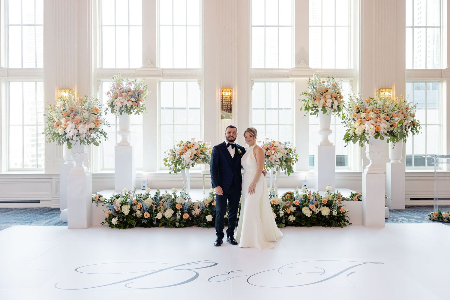 Elegant wedding ceremony setup in bright white venue with tall windows and floral arrangements along the aisle.