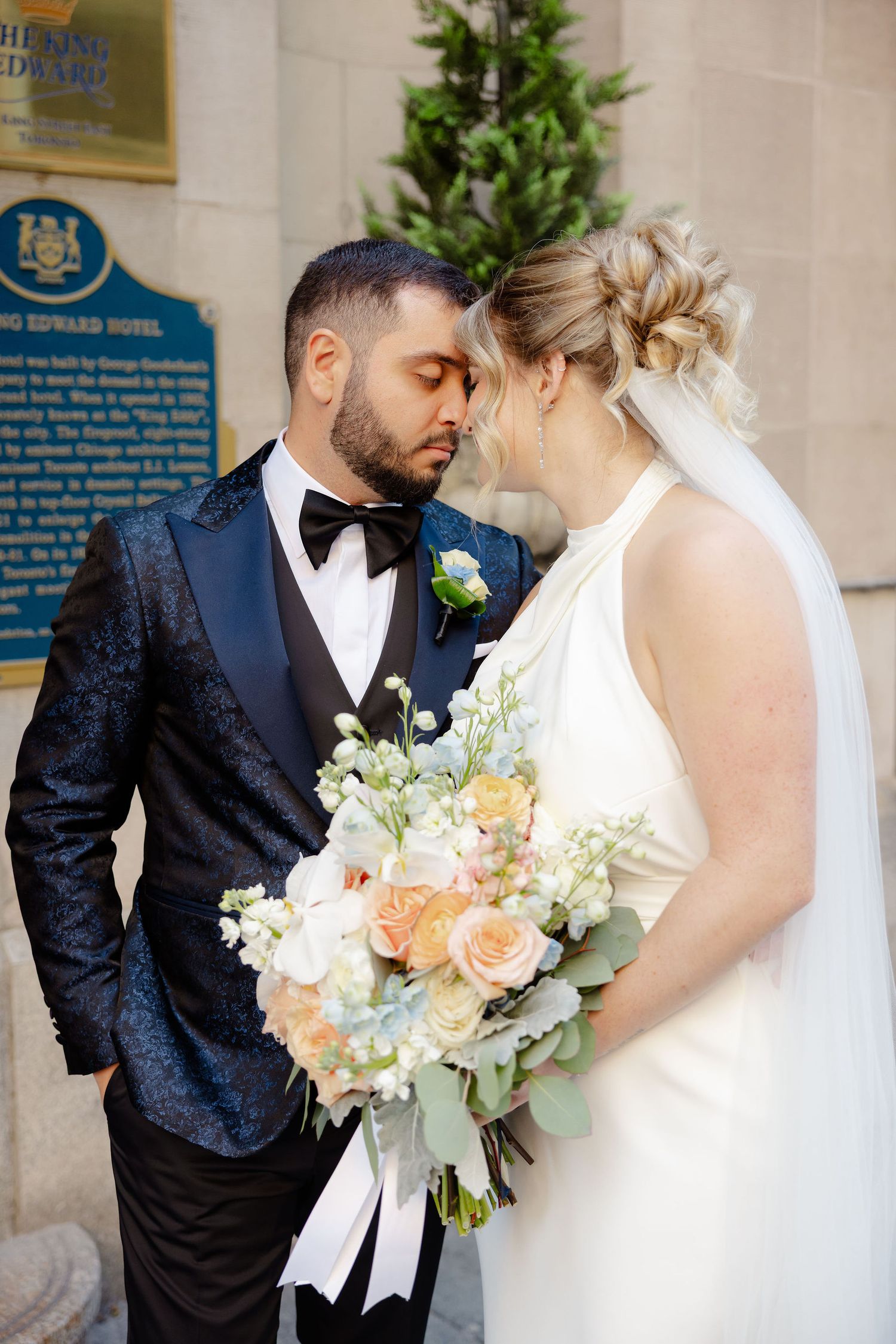 Romantic wedding couple sharing intimate moment with peach and white bouquet against elegant backdrop.