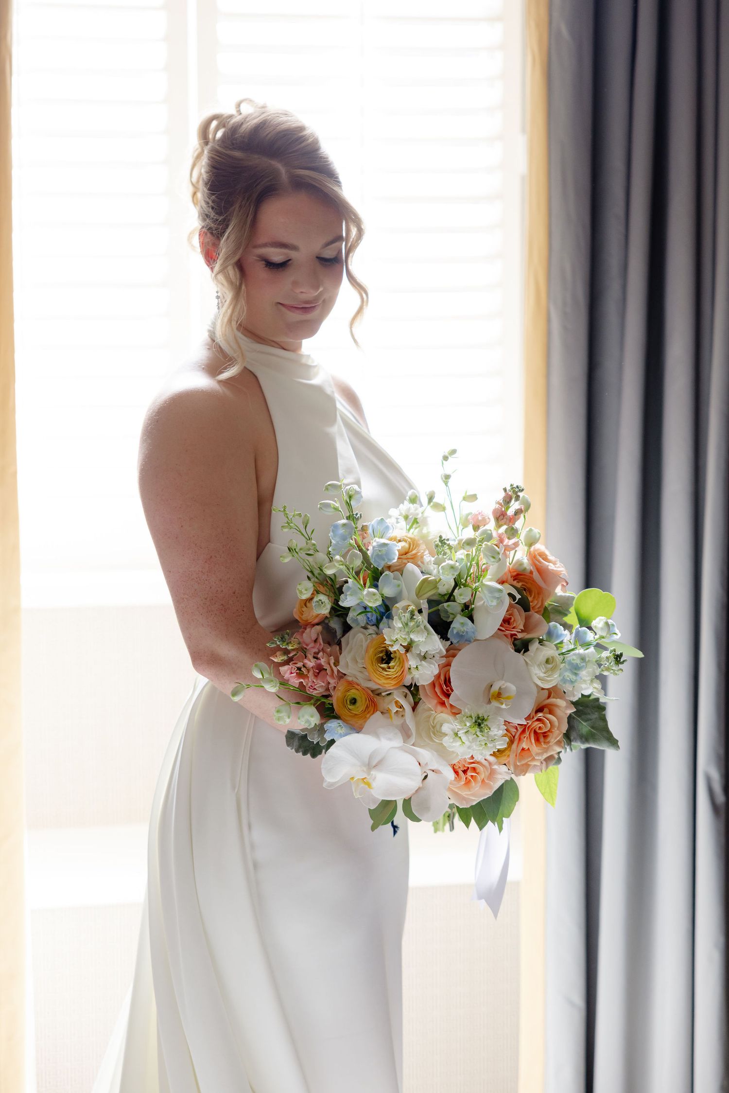 Elegant white wedding dress with a colorful bouquet of orange and white flowers against a window.