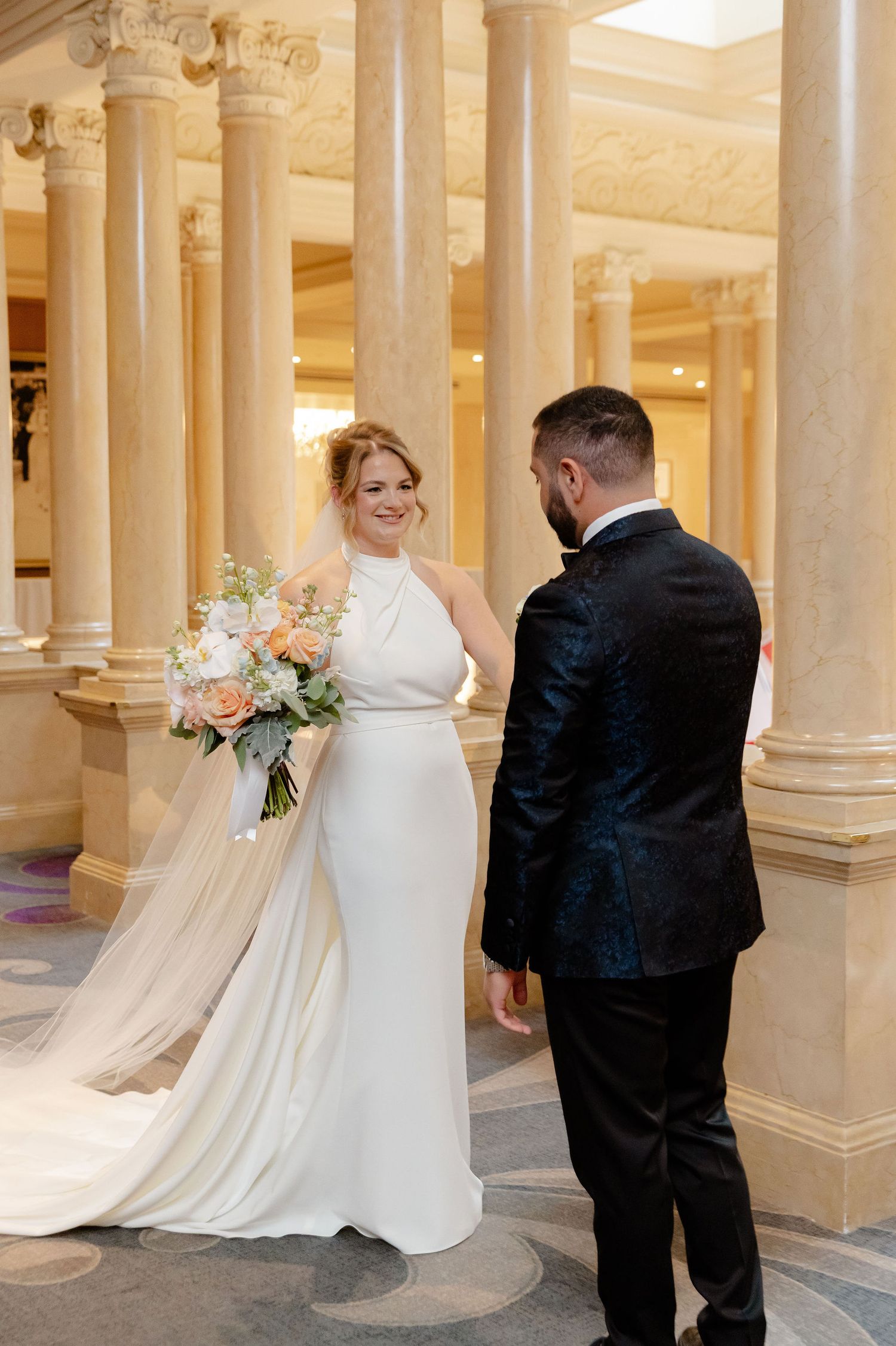 Elegant wedding couple walks through a columned hallway with soft lighting and flowing white dress trailing behind.