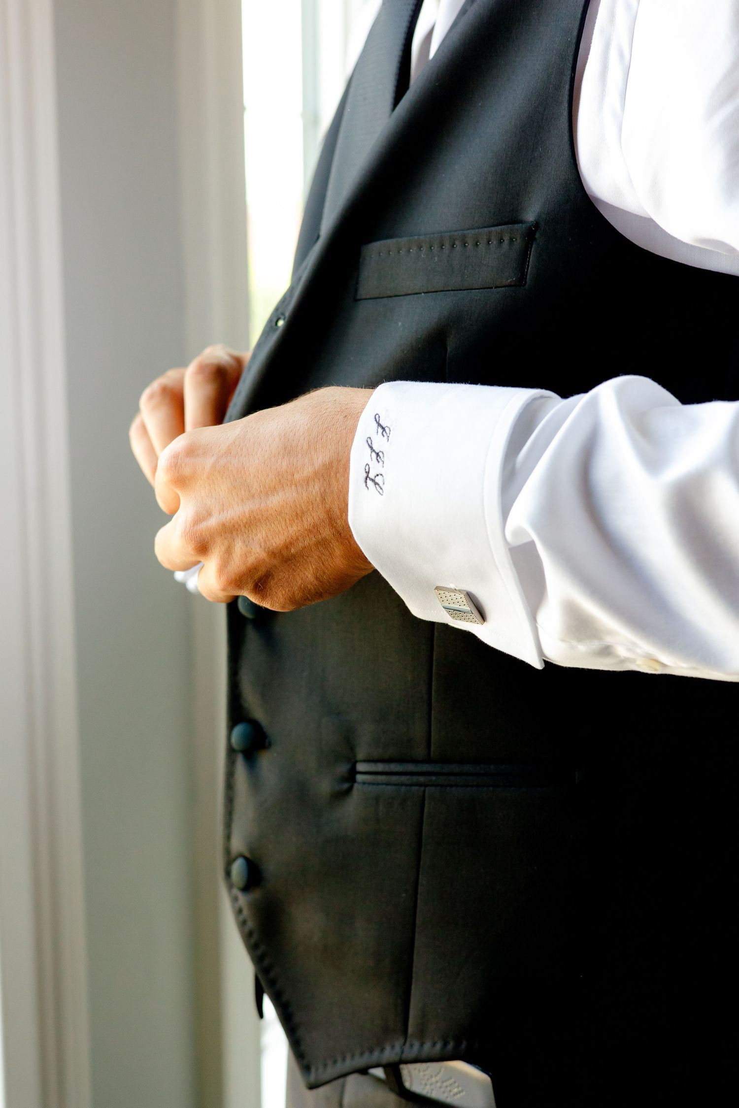 Close-up of hands adjusting a black formal vest with monogrammed shirt cuff visible.