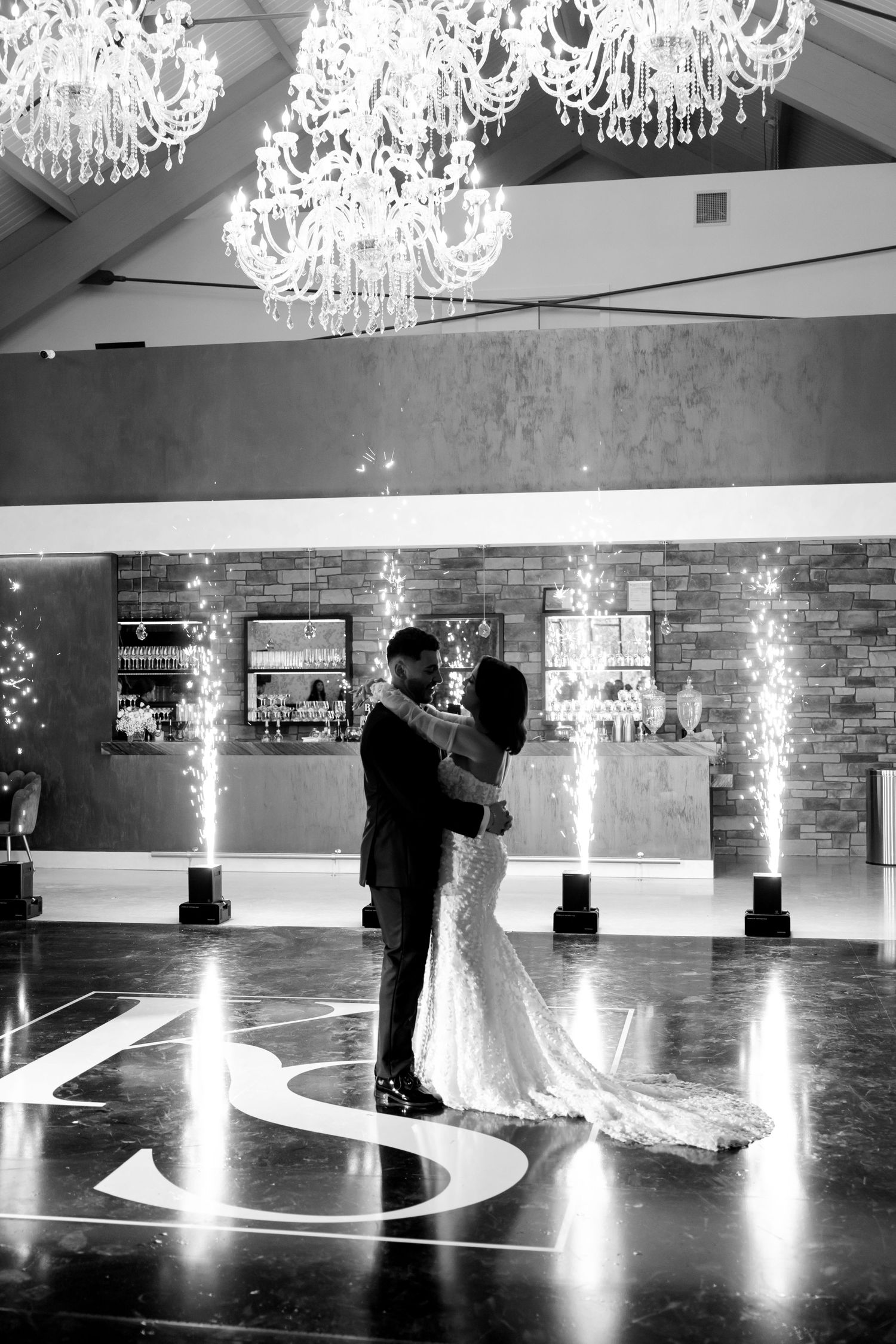 Wedding couple dances under elegant crystal chandeliers with lit monogram on floor at elegant reception venue.