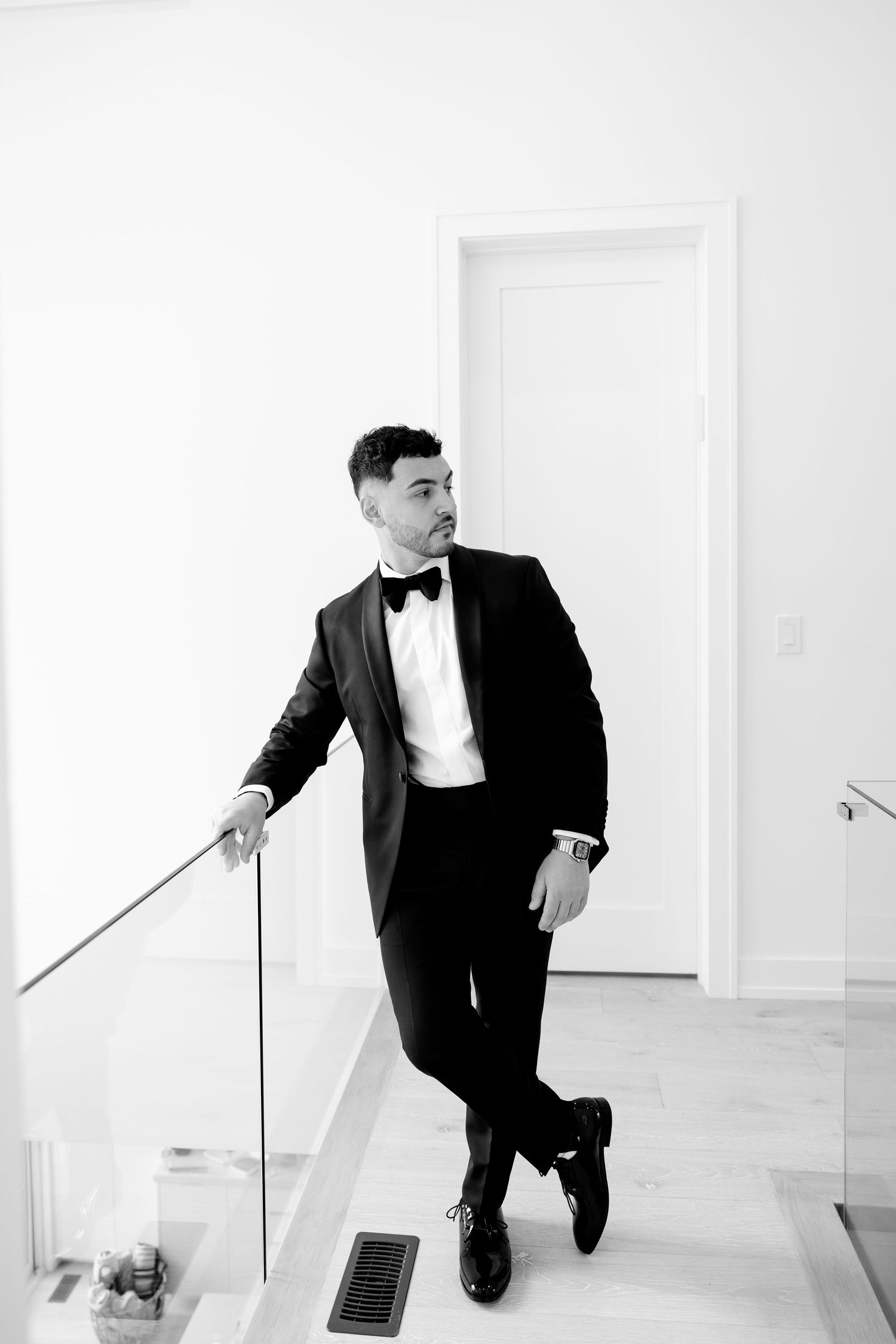 Black and white portrait of groom in tuxedo posing against glass railing and white walls.