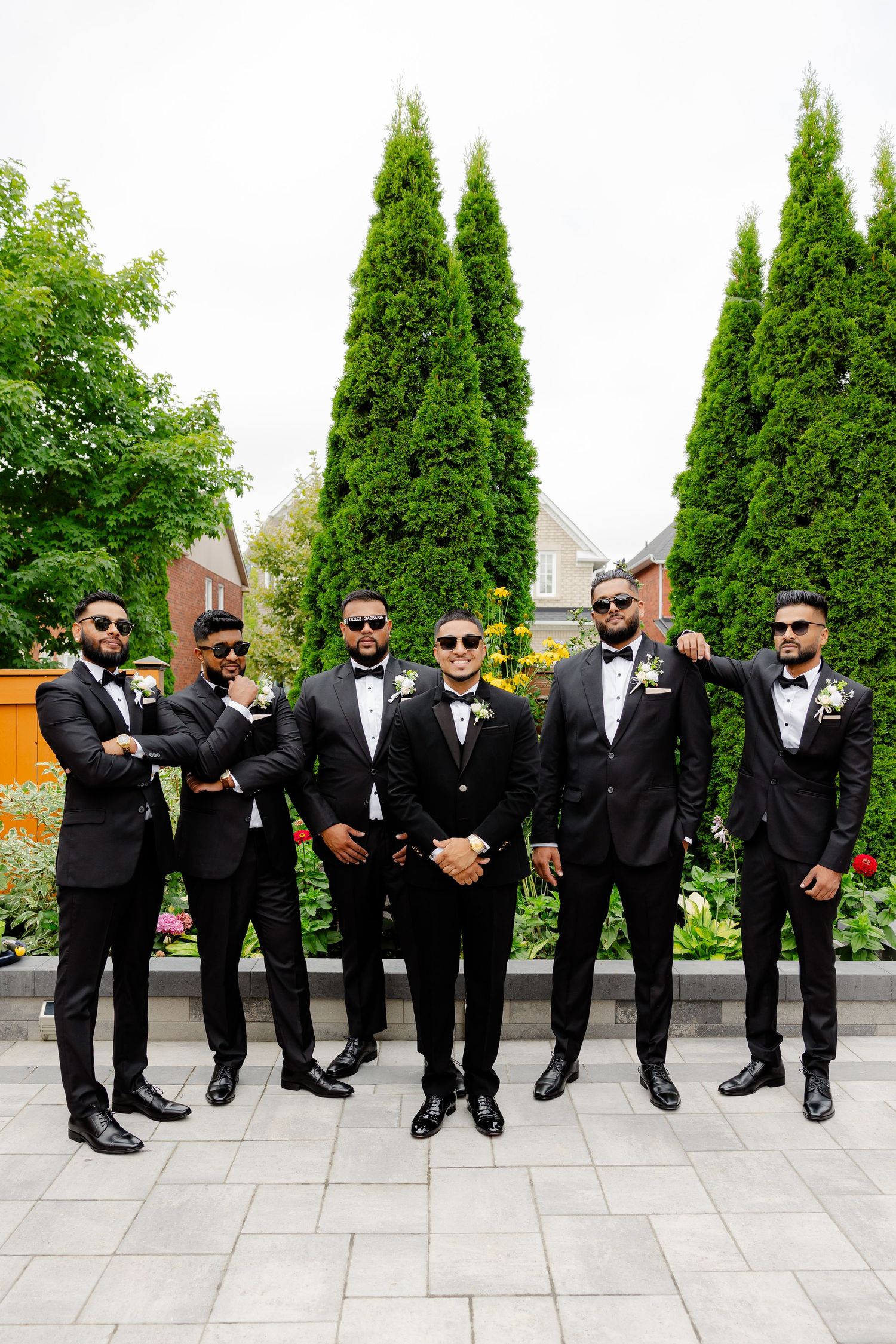 Groomsmen in matching black tuxedos and sunglasses pose together against tall cypress trees in a garden setting.