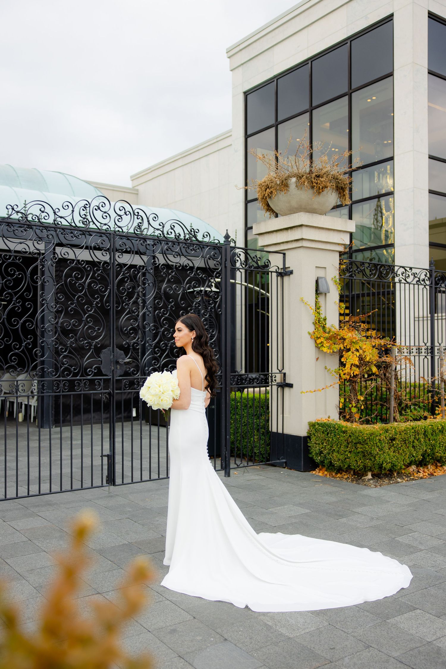Bride in flowing white gown holds bouquet while standing near decorative iron gates.