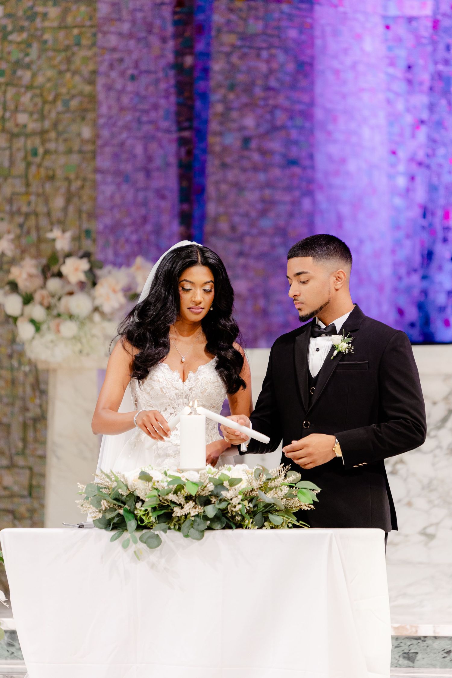 Newlyweds performing unity ceremony at decorated altar with white floral arrangements.