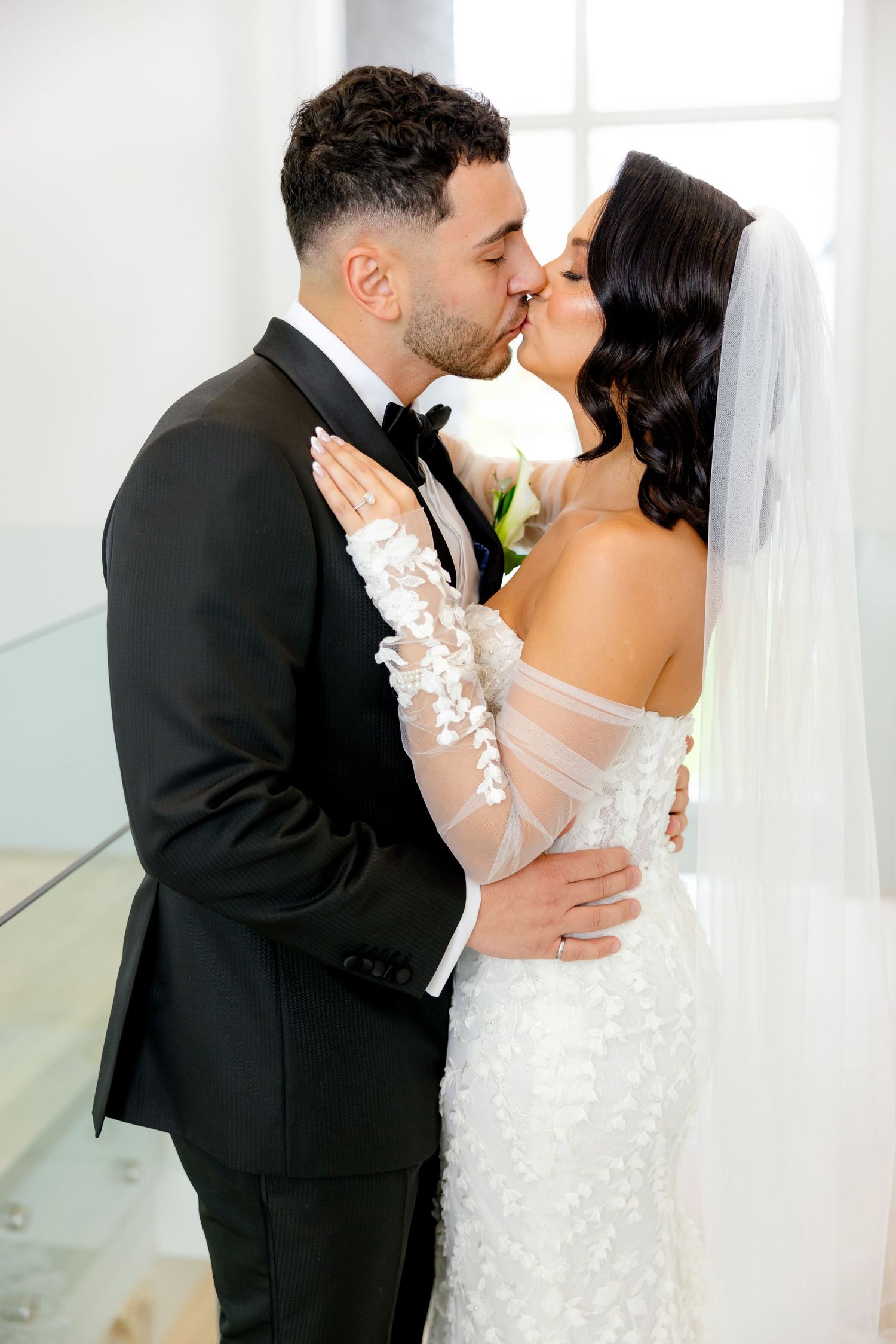 Newlyweds share intimate kiss in bright white room during wedding photoshoot.