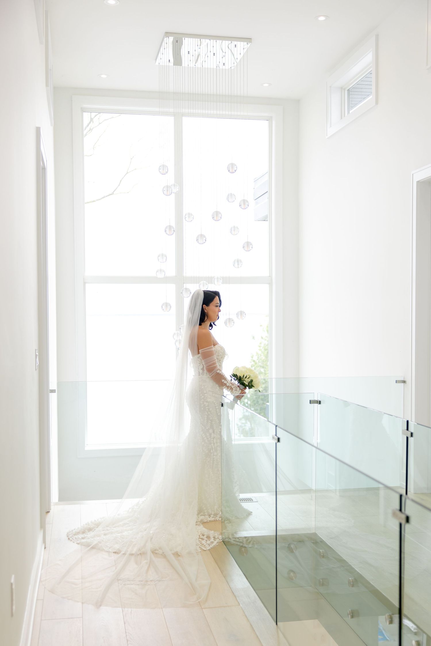Bride in fitted lace gown poses by large window in modern white interior with glass railings.