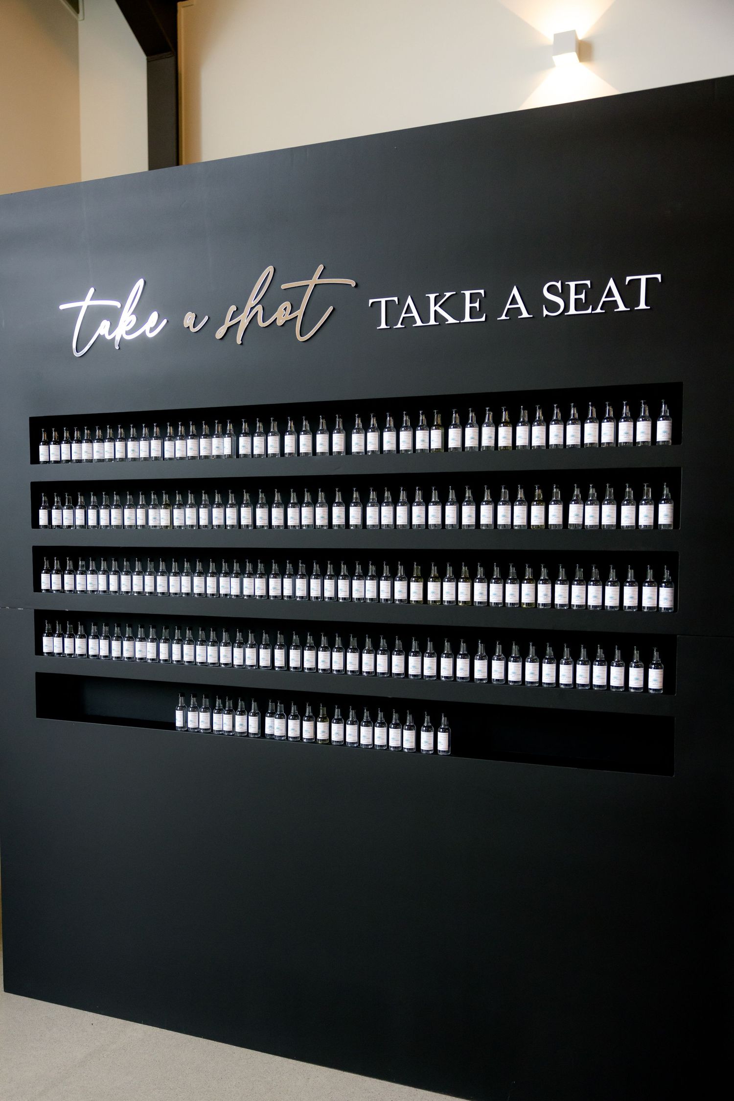 Black wall display with illuminated bottles arranged in rows and 'Take a Shot, Take a Seat' text at the top.