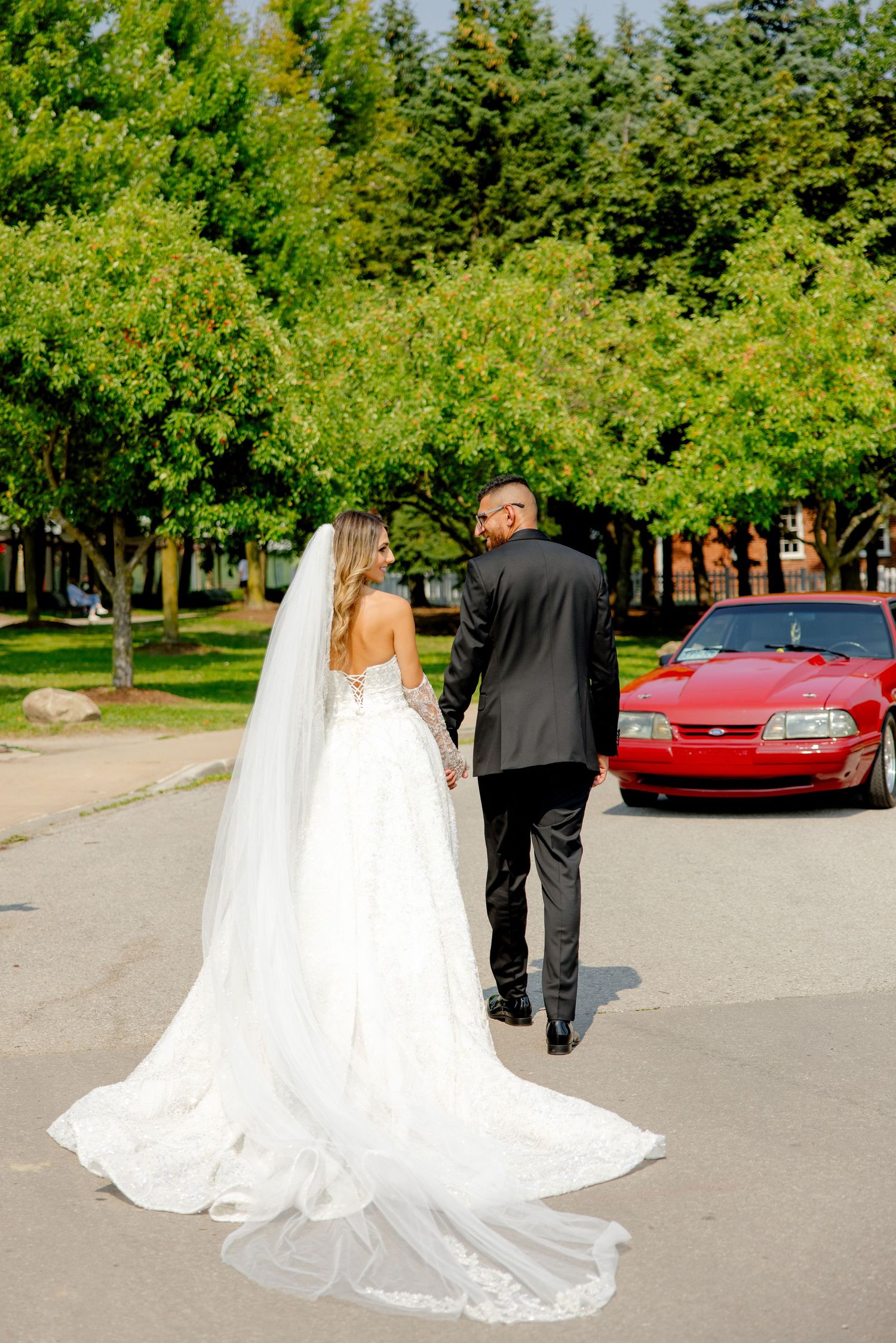 Bride in flowing white dress and groom walk away from camera with red car in background.
