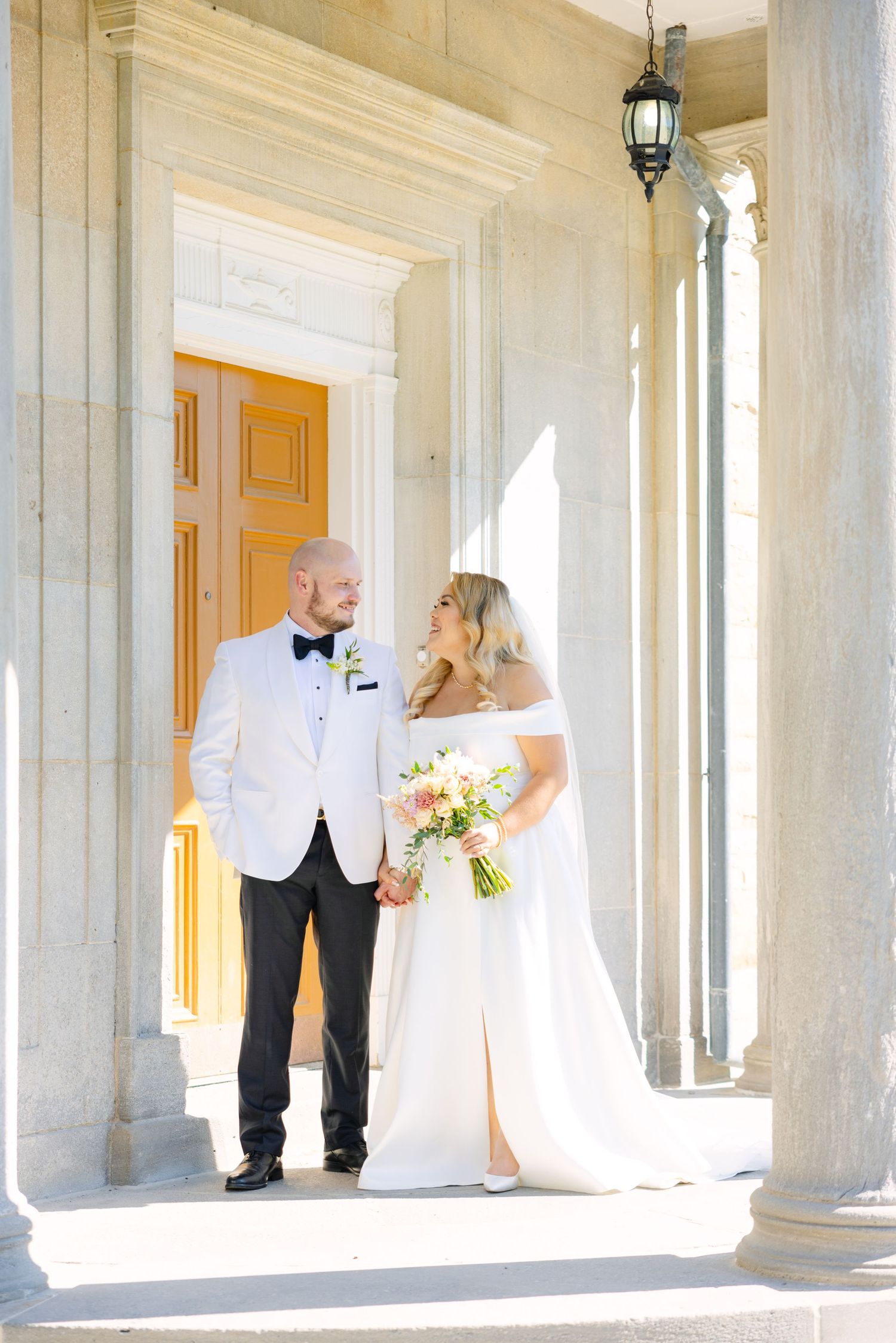 A wedding couple shares a joyful moment in a sun-lit portico with elegant columns and an orange door.