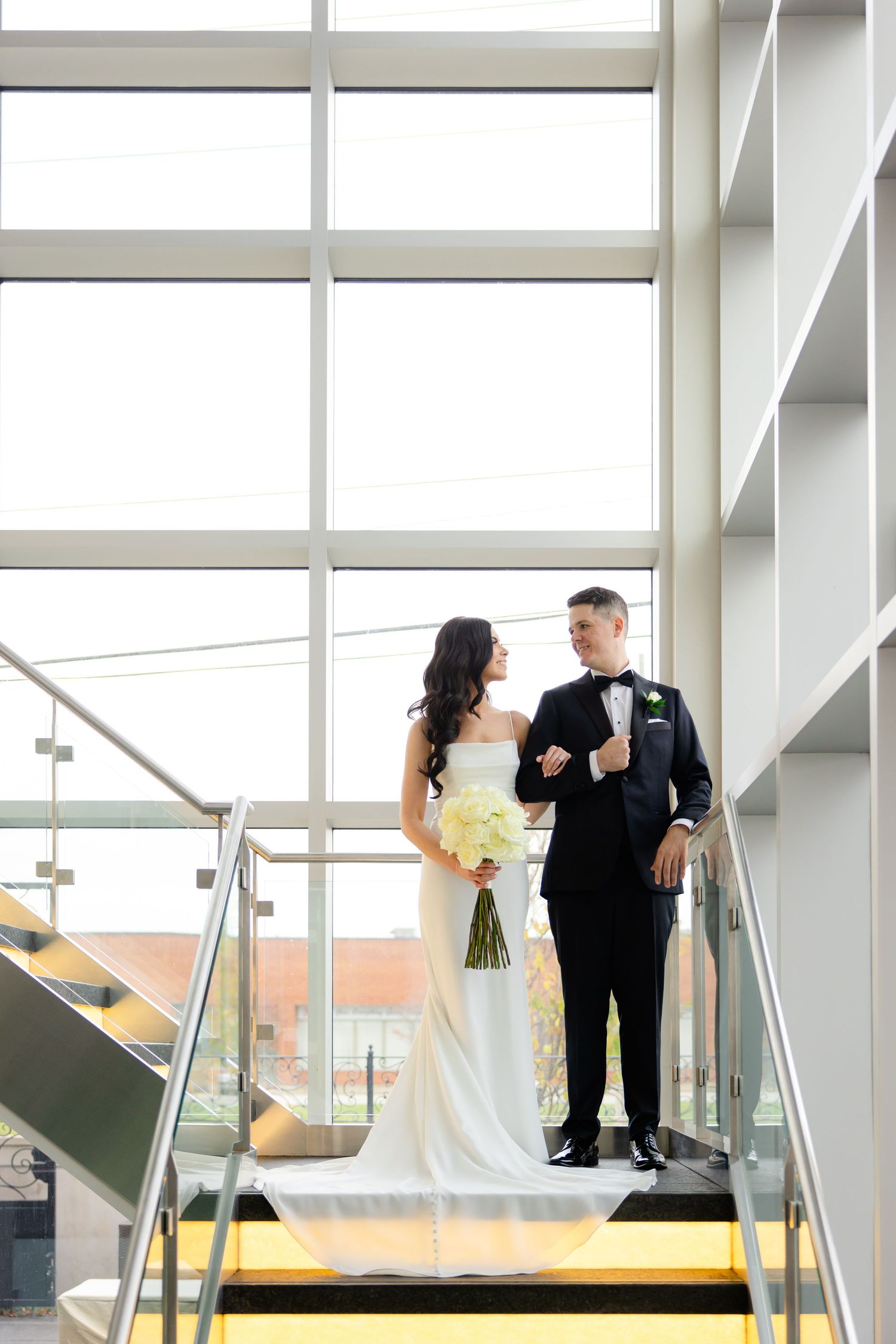 Couple in formal attire share intimate moment on modern staircase with natural light.