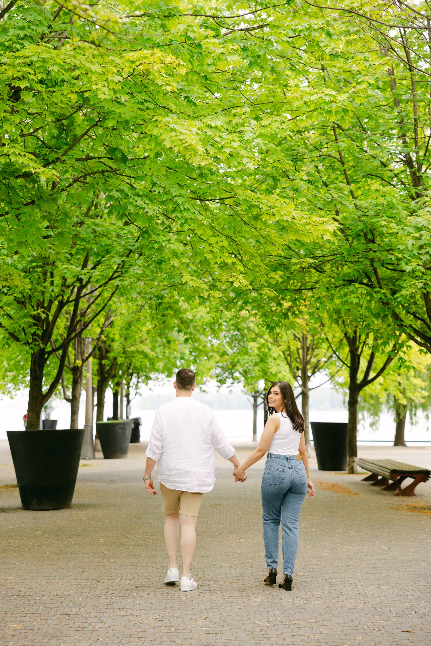 A couple strolls together under vibrant green trees along a brick pathway.