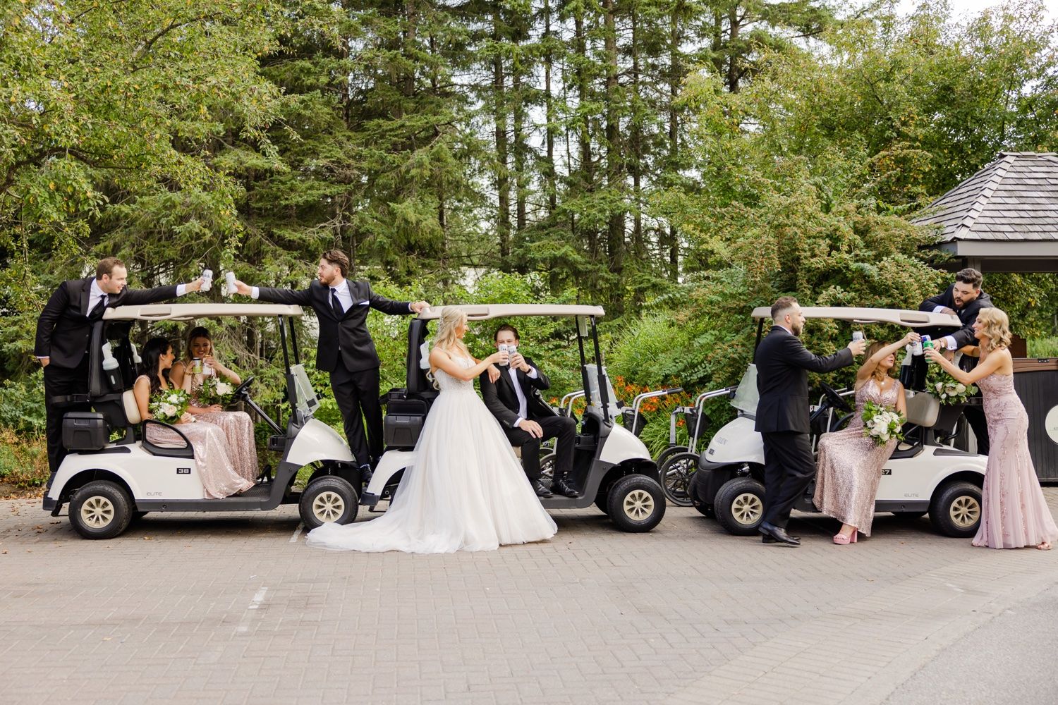 Wedding party gathered around white golf carts at an outdoor venue surrounded by pine trees.