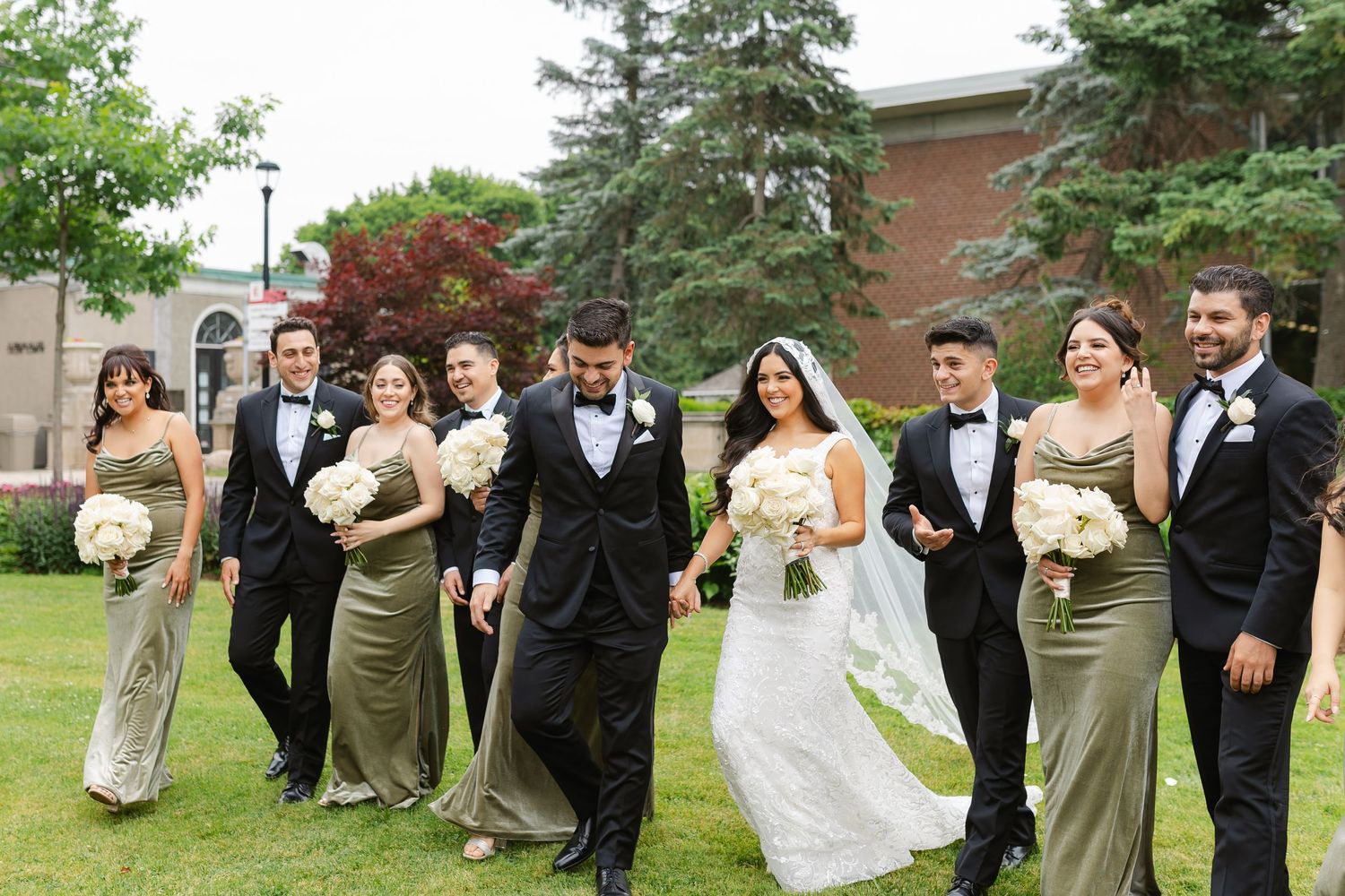Wedding party walking in formation wearing sage green dresses and black tuxedos.