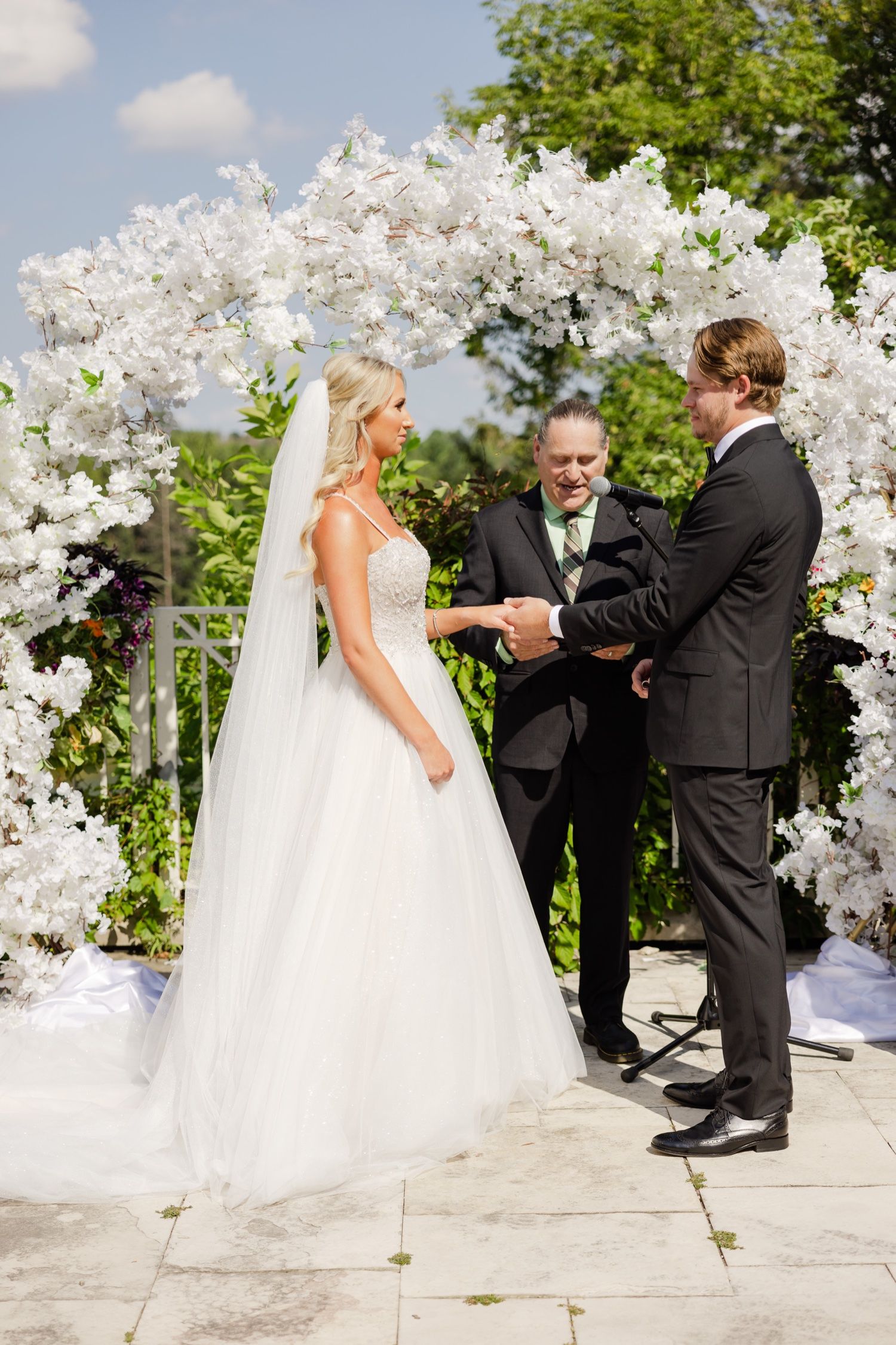 Wedding ceremony taking place under a stunning white floral arch on a stone patio.