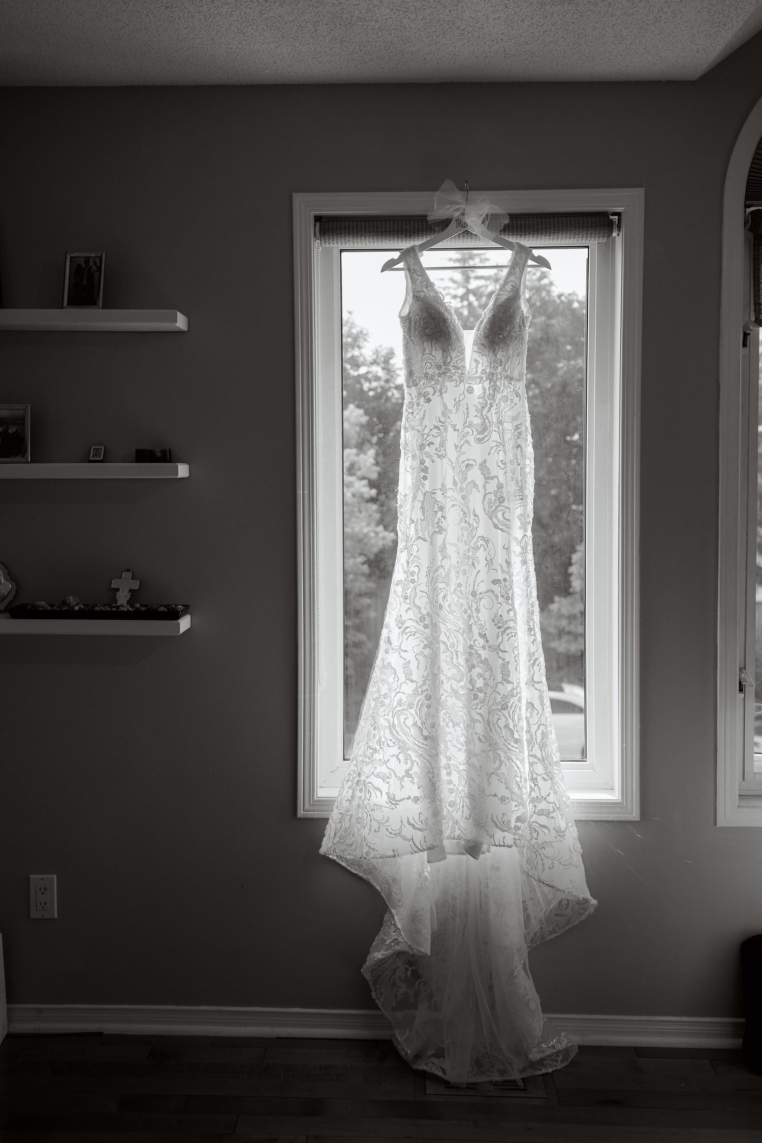 Lace wedding dress hanging in window with natural light creating silhouette.