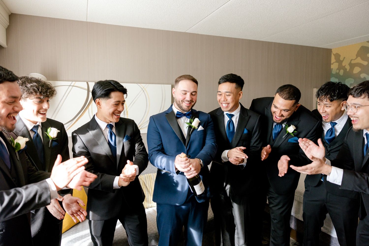 Groomsmen gathered in formal attire sharing pre-wedding laughs in a hotel room.