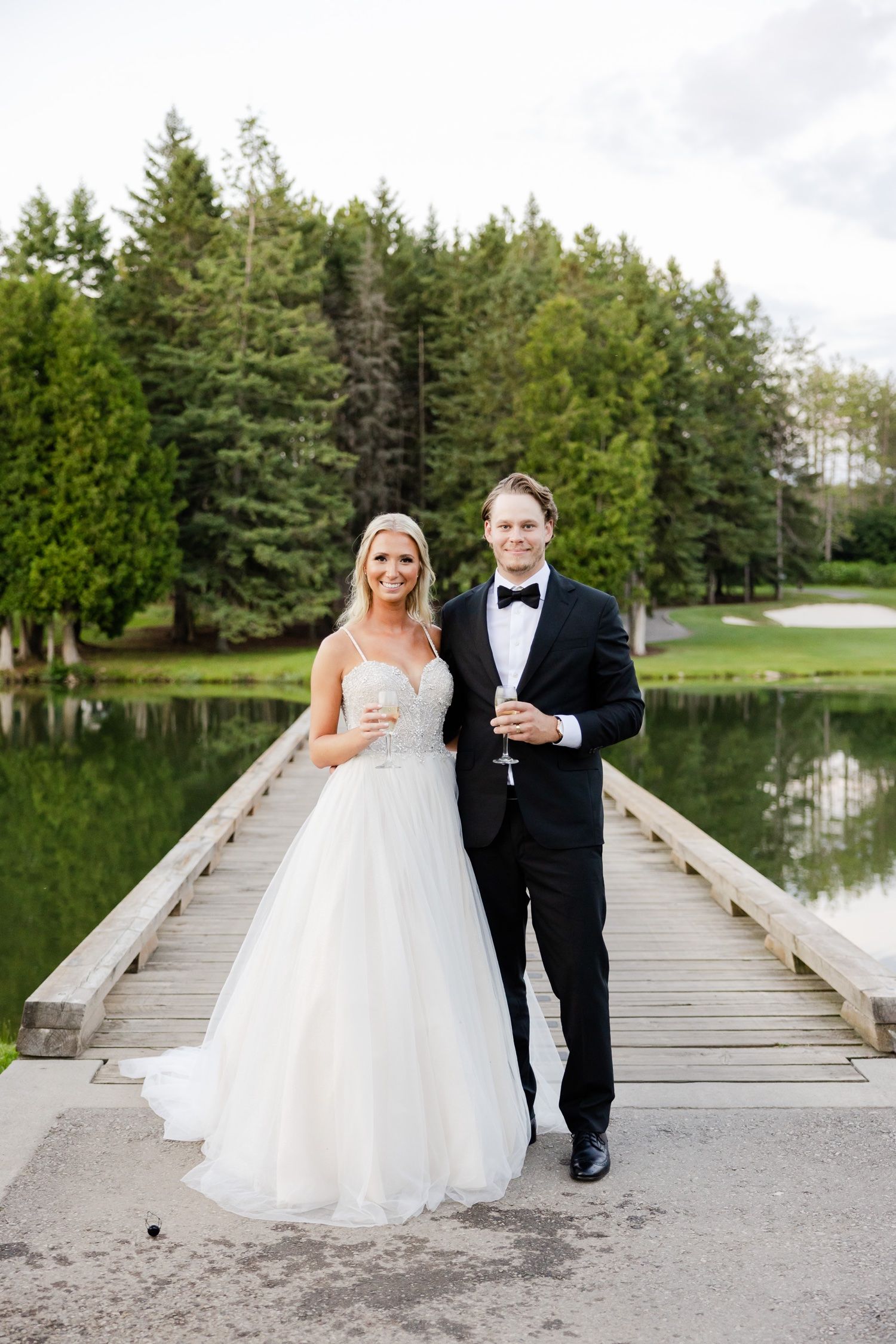A newlywed couple stands on a wooden dock surrounded by pine trees and a serene lake.