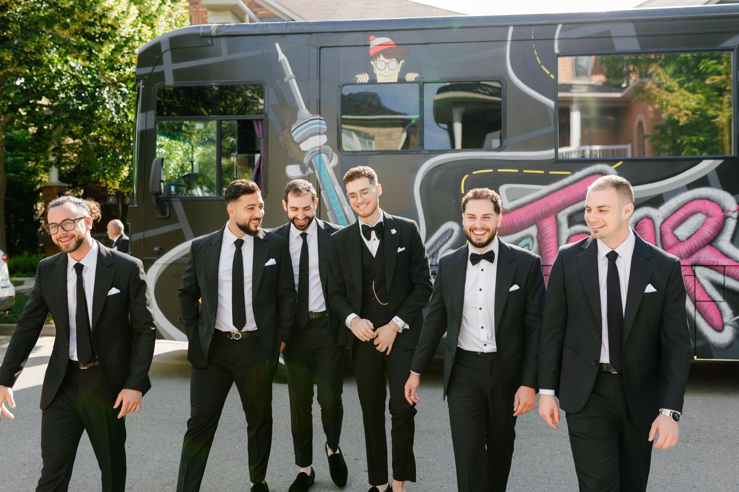 Groomsmen in black suits and ties pose together in front of a vintage vehicle on a sunny day.