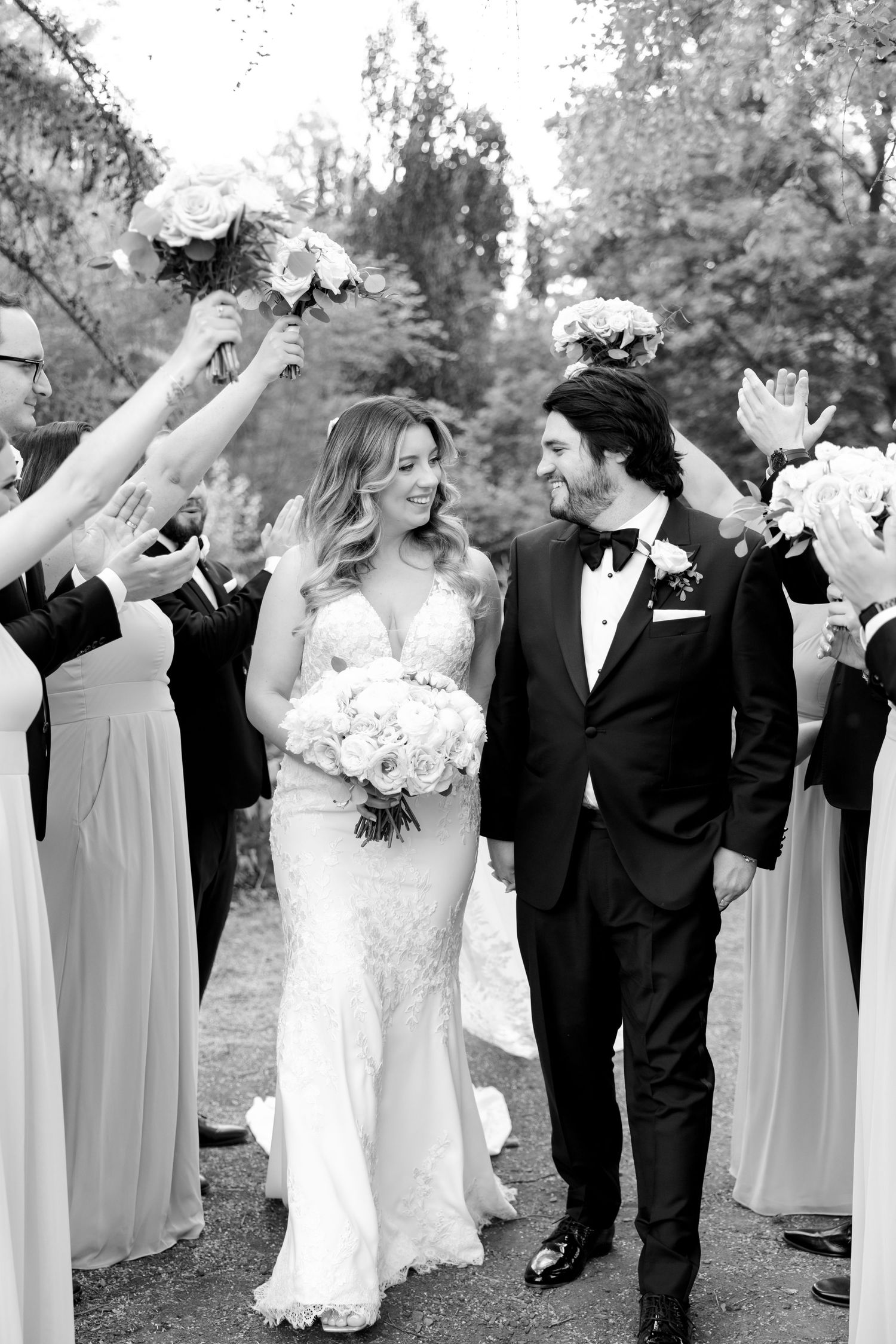 Black and white photo of bridal party raising bouquets as couple walks through garden path.