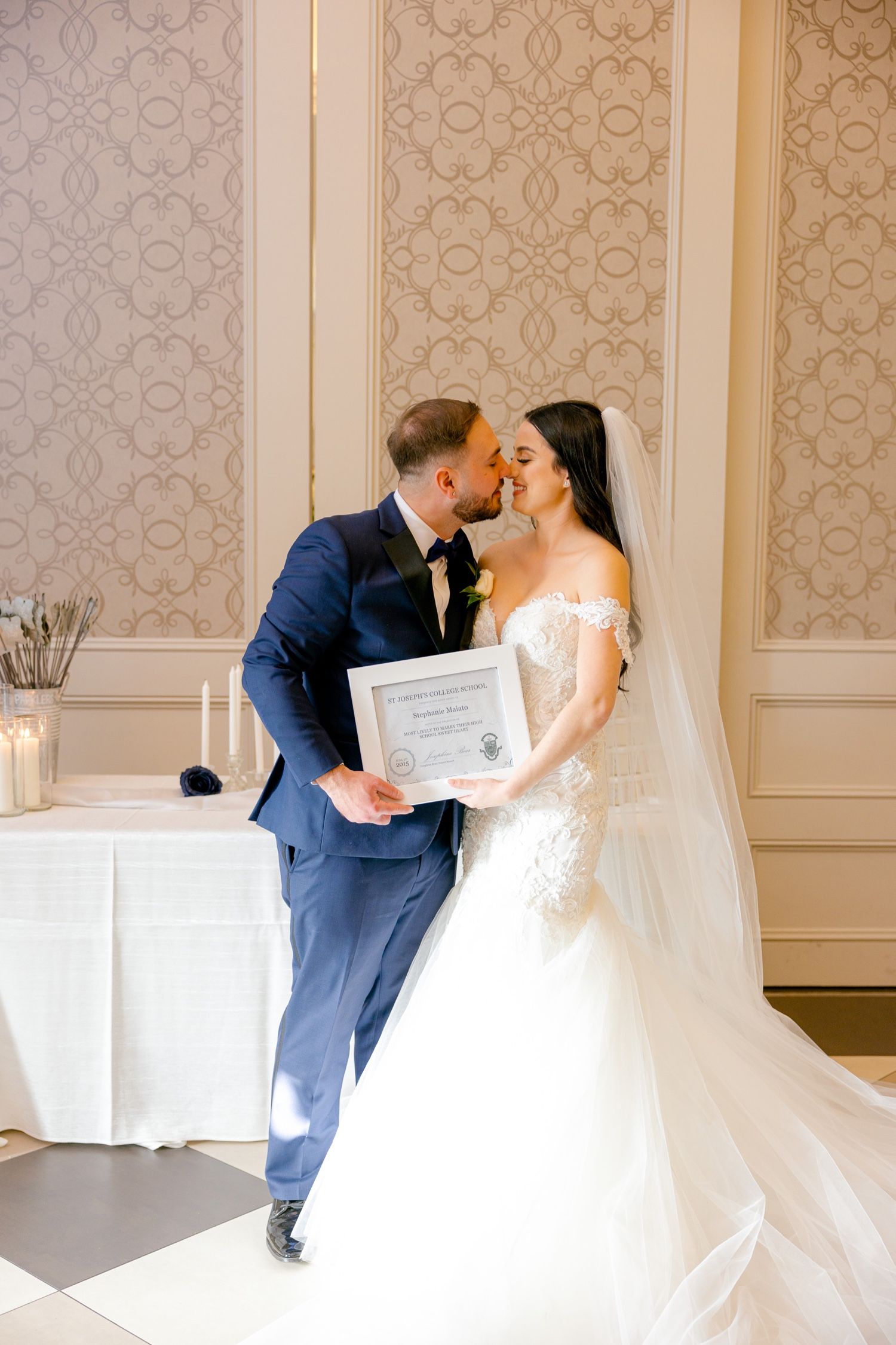 Elegant wedding couple posing with certificate in front of patterned wallpaper.