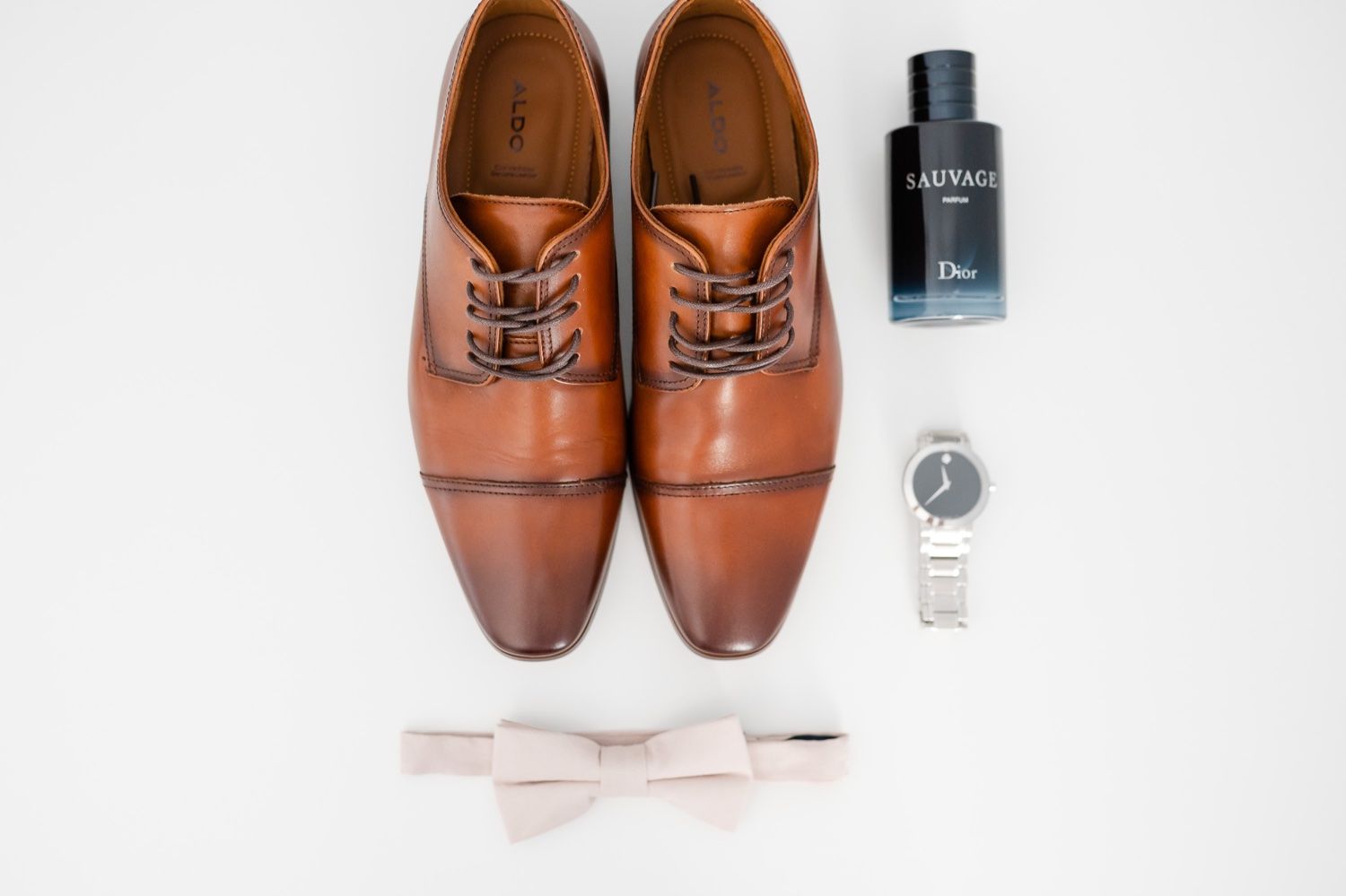 Brown leather dress shoes with bow tie and cologne bottle arranged on white surface.
