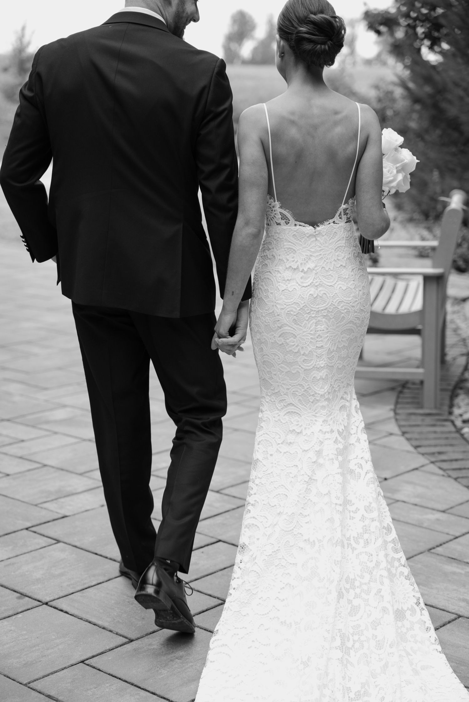 Back view of a couple in wedding attire walking hand in hand on stone pavers.