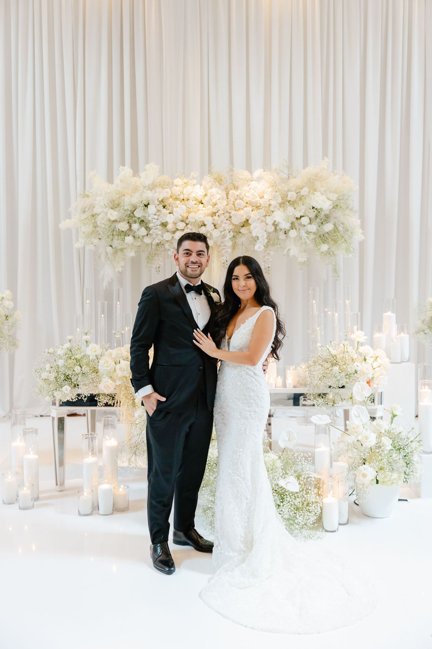 Wedding couple posing under luxurious white floral arch with candlelit decor.