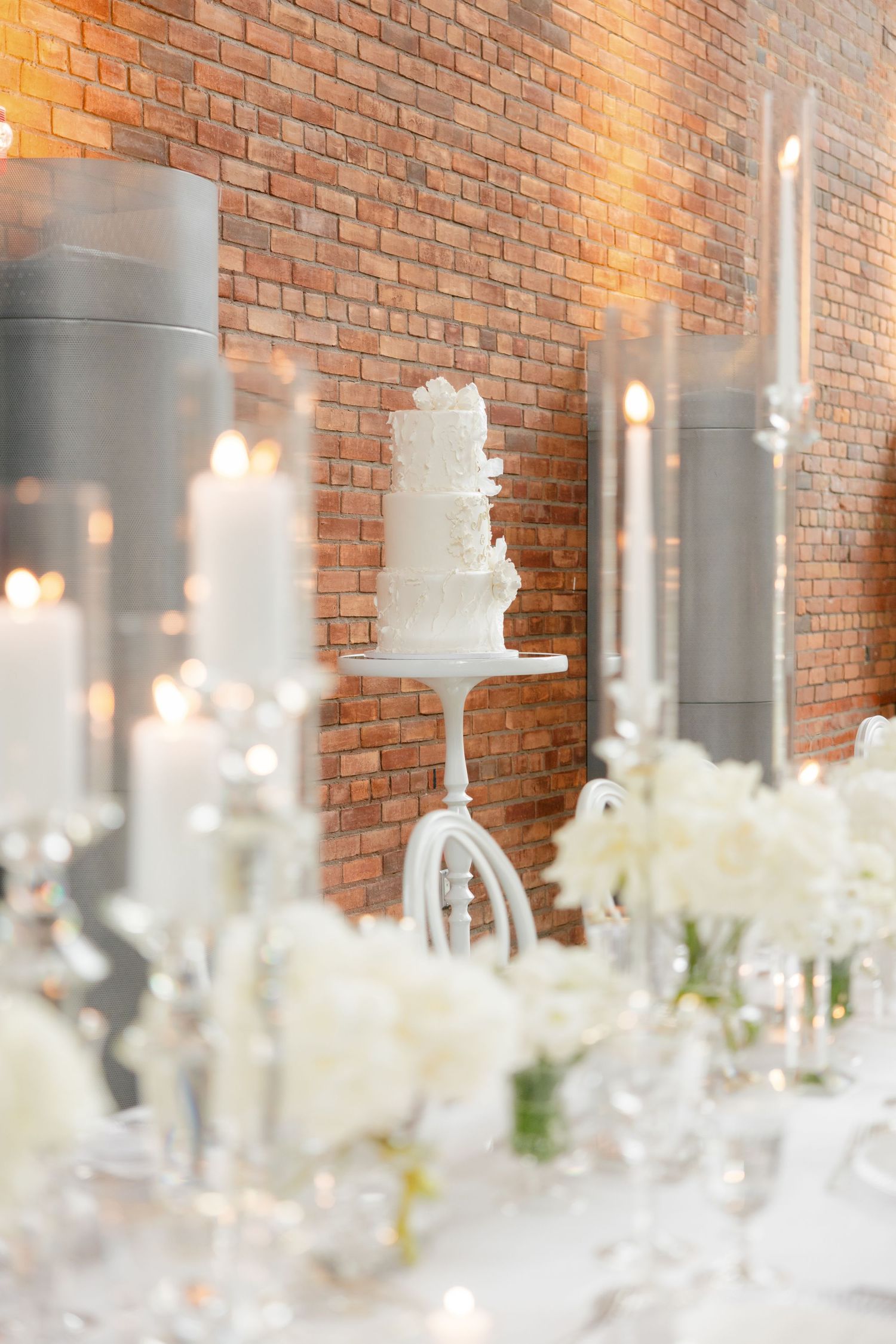 White tiered wedding cake displayed against exposed brick wall with glass candle holders and floral decor.