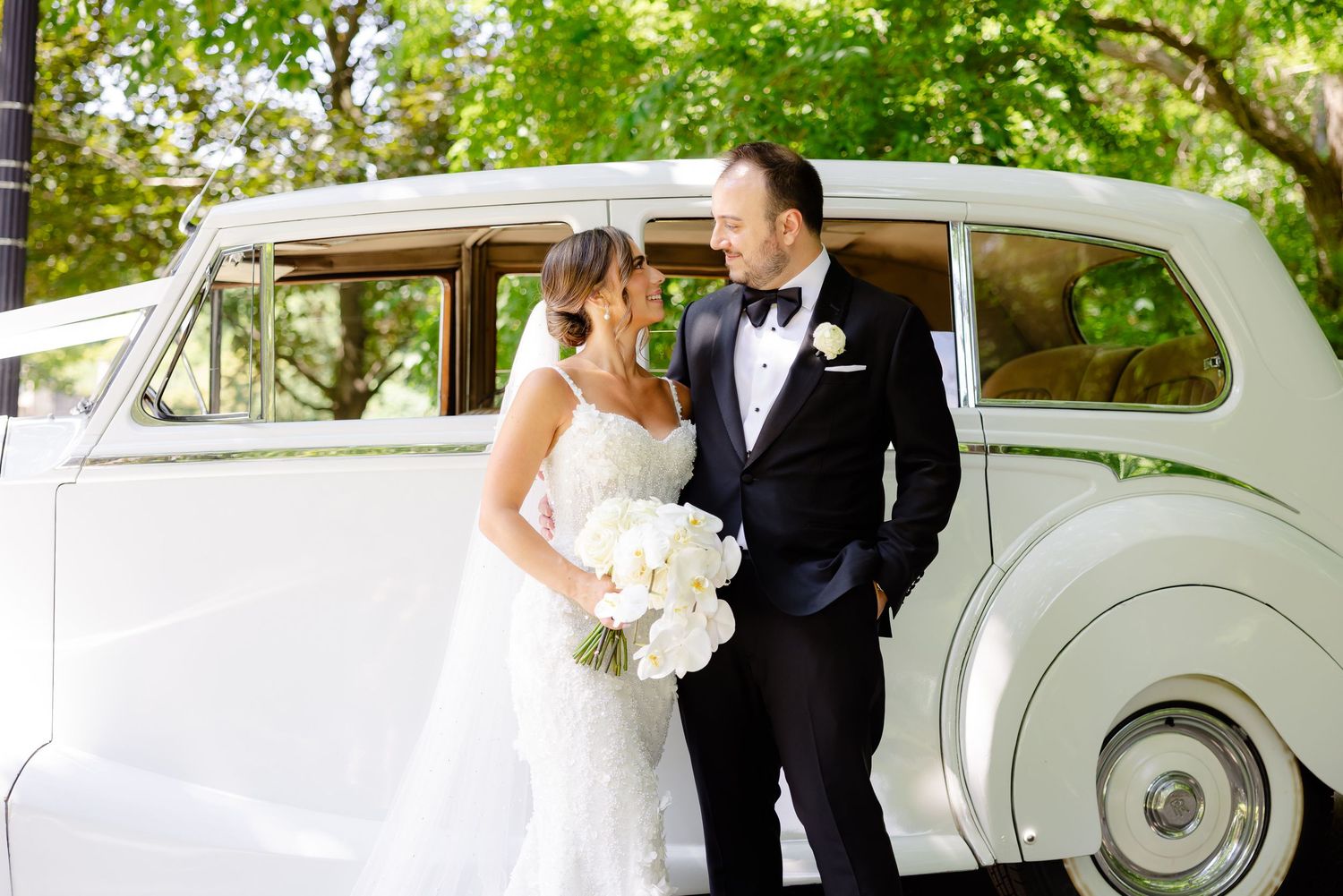 Elegant couple poses by vintage white Rolls Royce at wedding with white bouquet and classic formal attire.