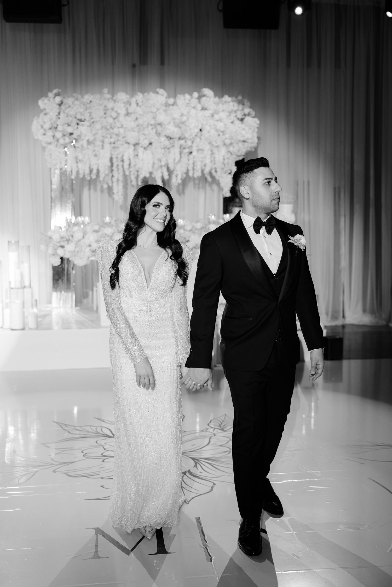 Black and white wedding photo of couple walking beneath an ornate floral arch.