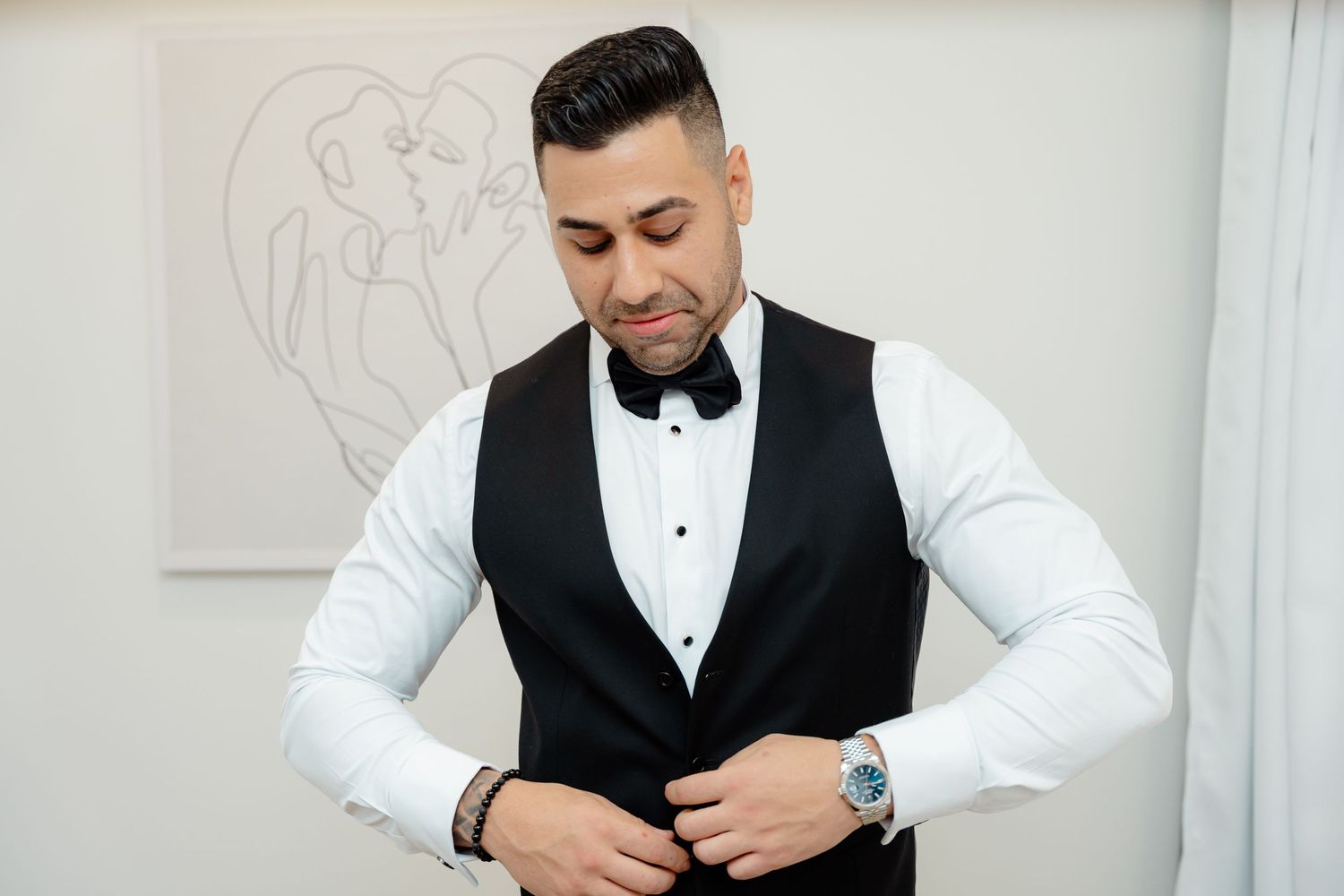 Groomsman in white shirt and black vest buttoning up formal attire.