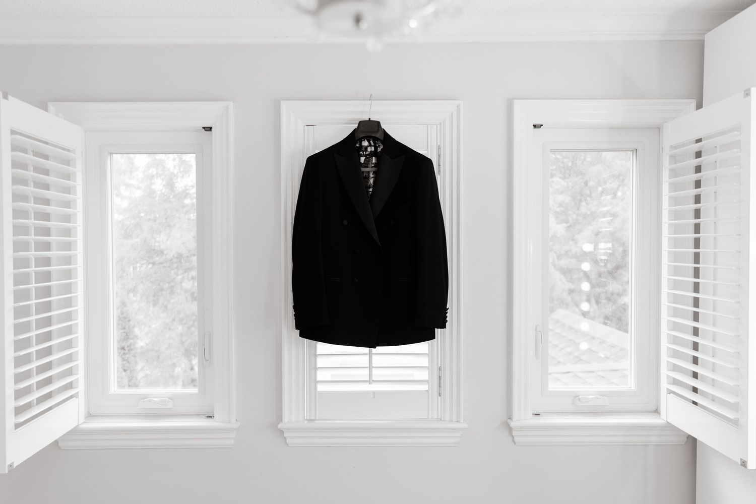 A minimalist white walk-in closet with clean lines features a hanging black suit jacket and louvered windows.