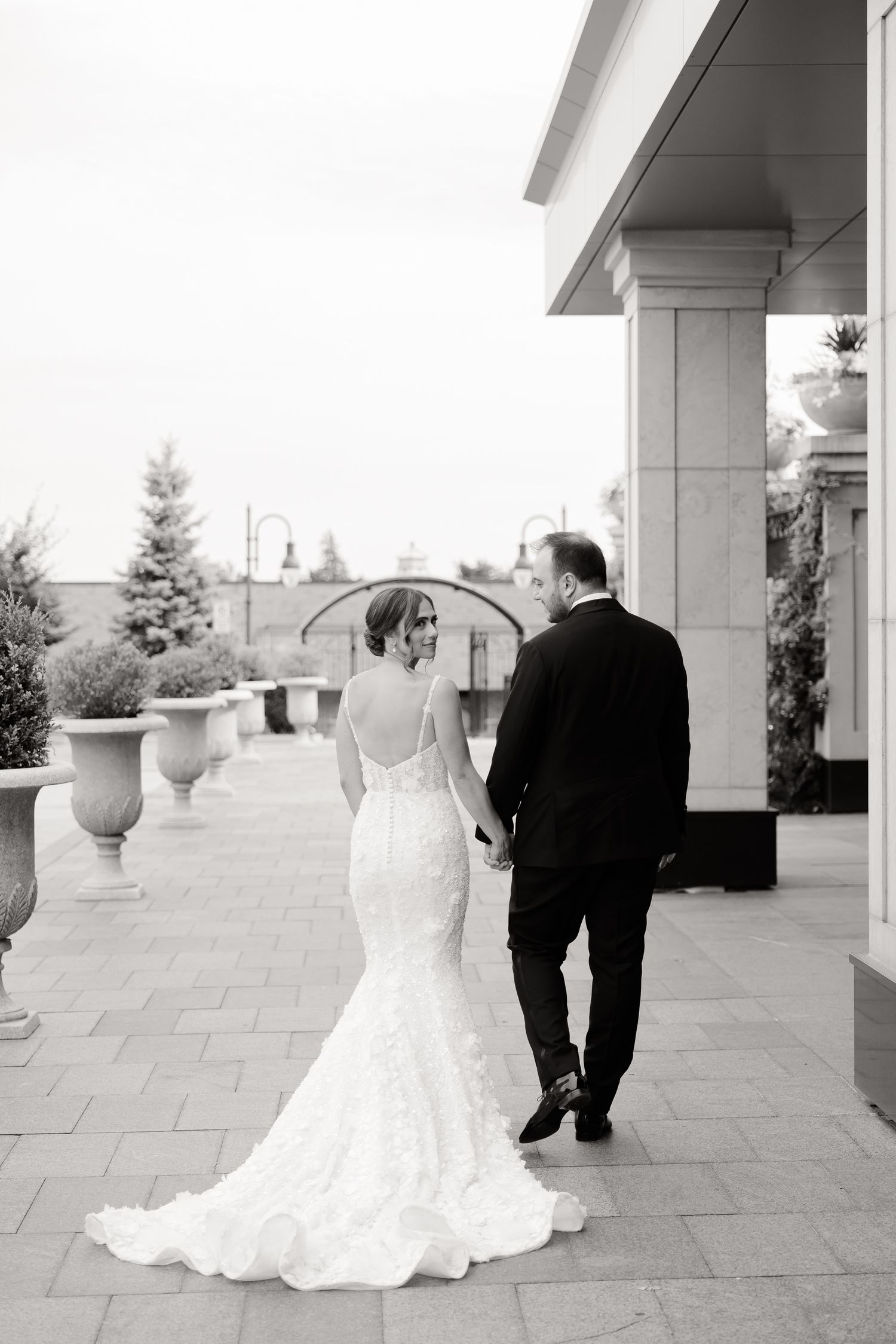 Black and white wedding photo with couple walking on stone terrace with elegant columns and balustrades.