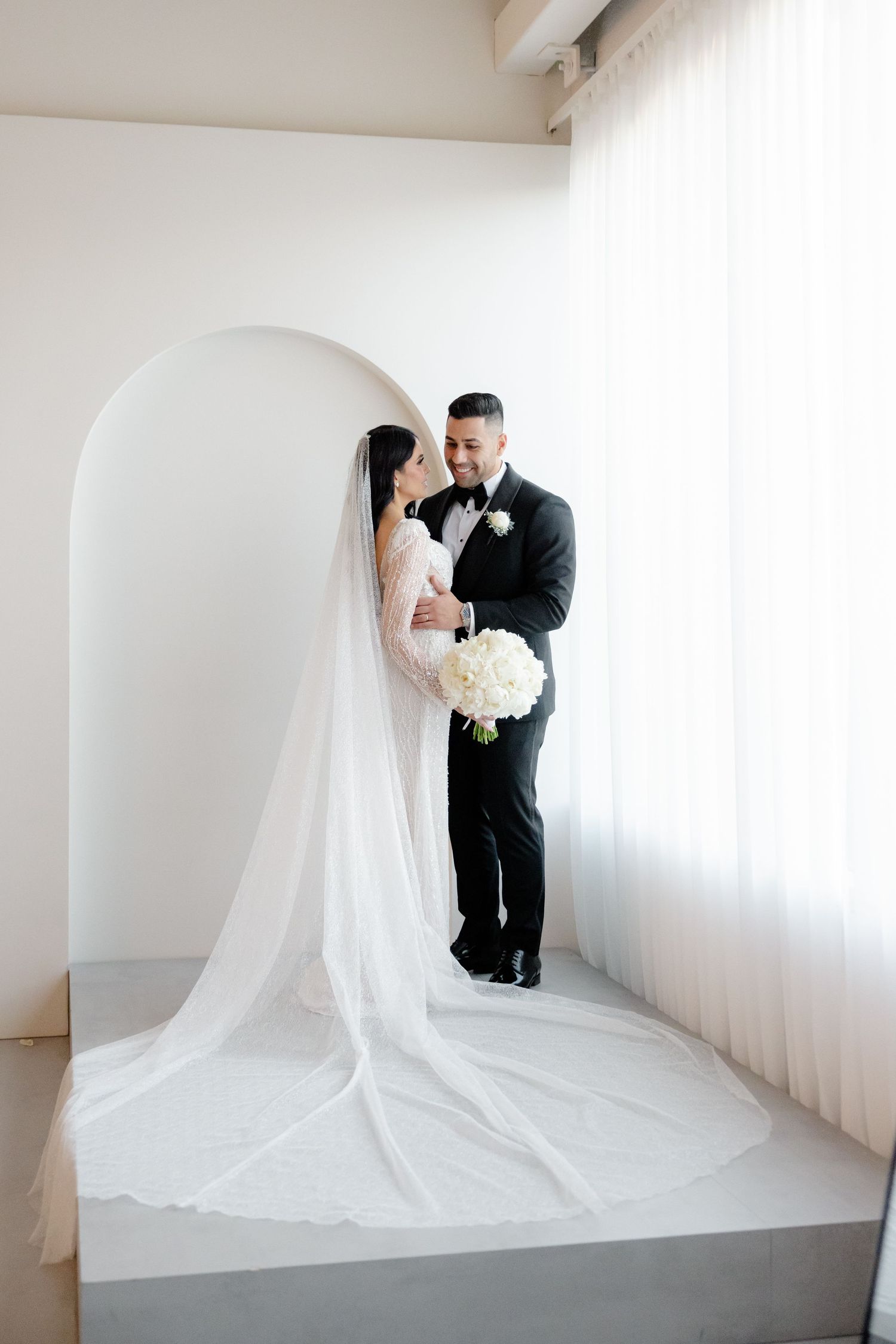 A wedding couple stands together under a white archway with a dramatic cathedral veil trailing behind.