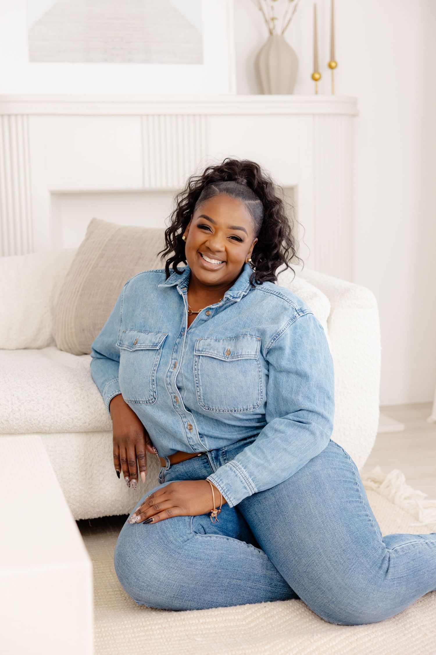 Someone wearing casual double denim outfit sits comfortably and smiles while relaxing on a white couch in a bright room.