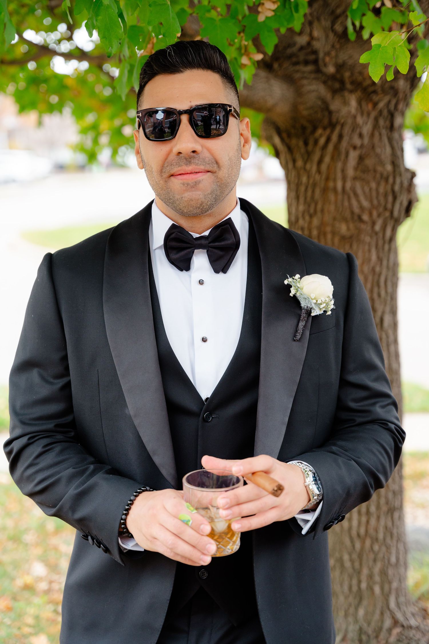 Dapper groom in black tuxedo and sunglasses holding a drink under a tree.