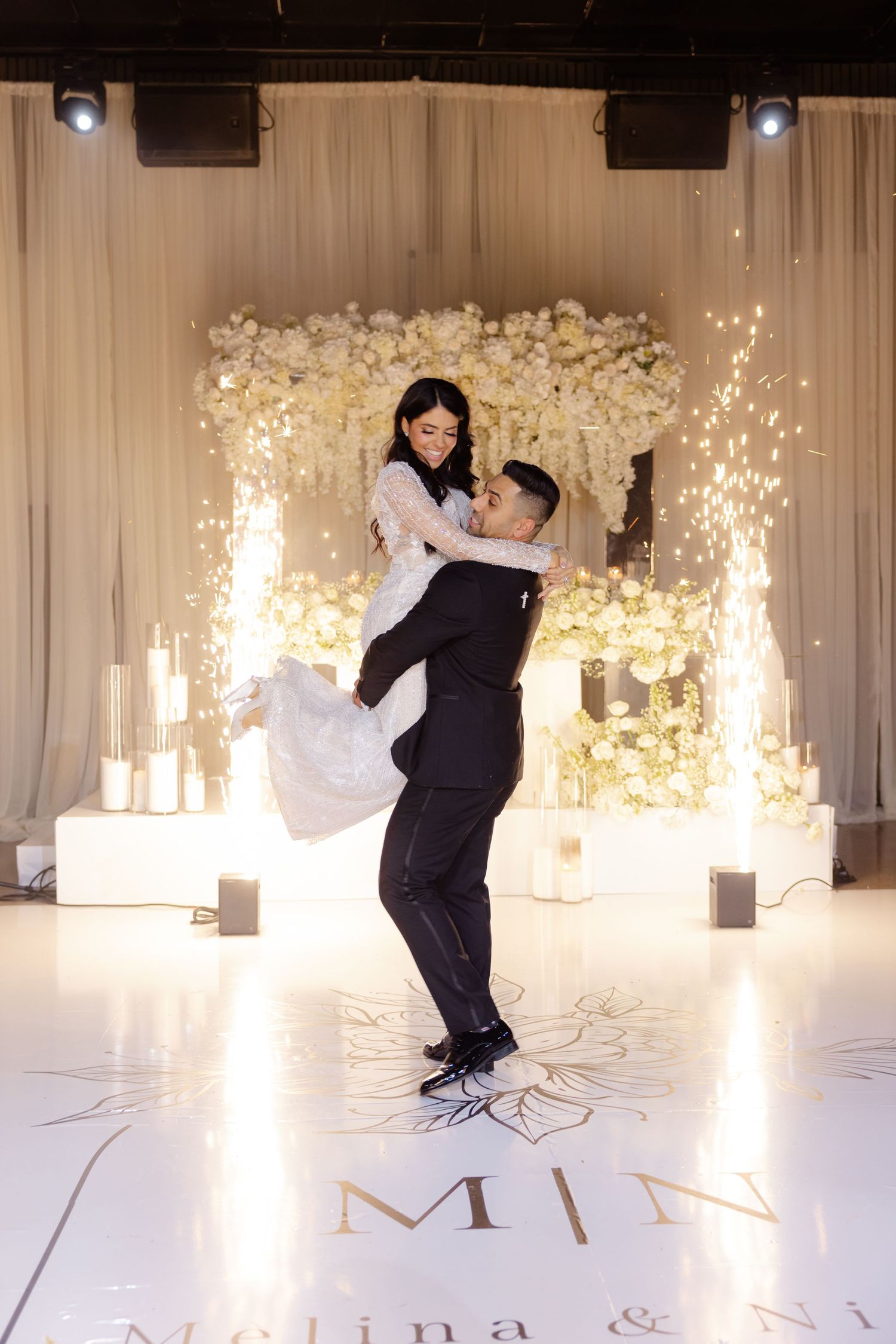 Newlyweds share a romantic dance moment with sparkler effects against a floral backdrop and monogrammed dance floor.