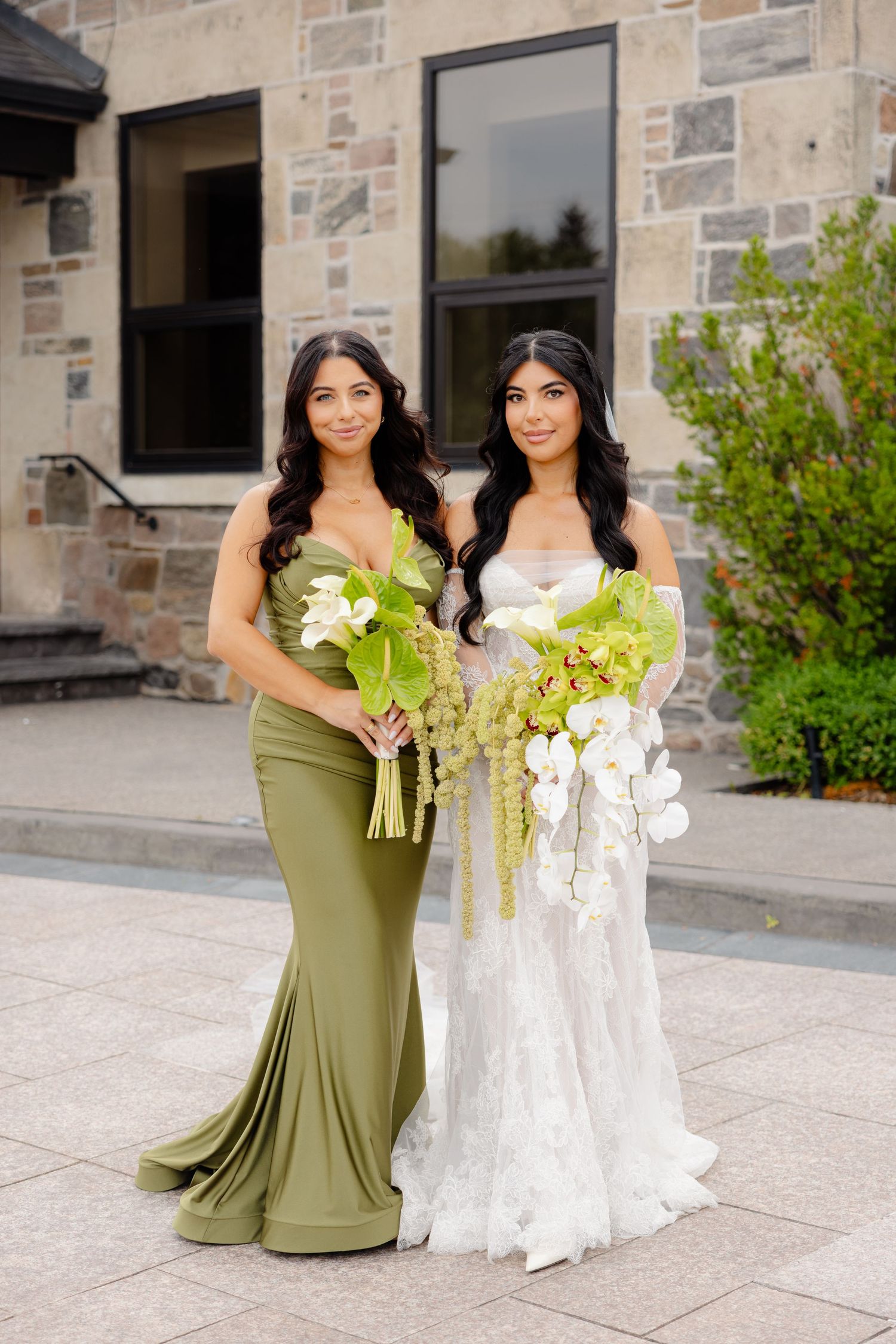 Two bridesmaids pose together on stone patio holding cascading white and green floral bouquets.