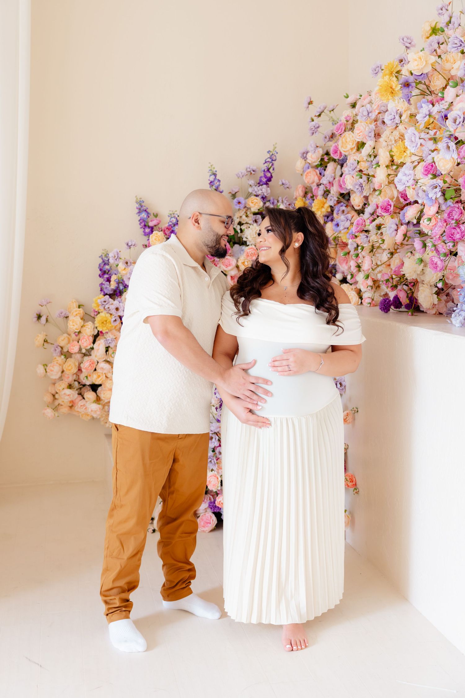 Romantic maternity photo with a couple in white outfits embracing in front of a floral wall backdrop.