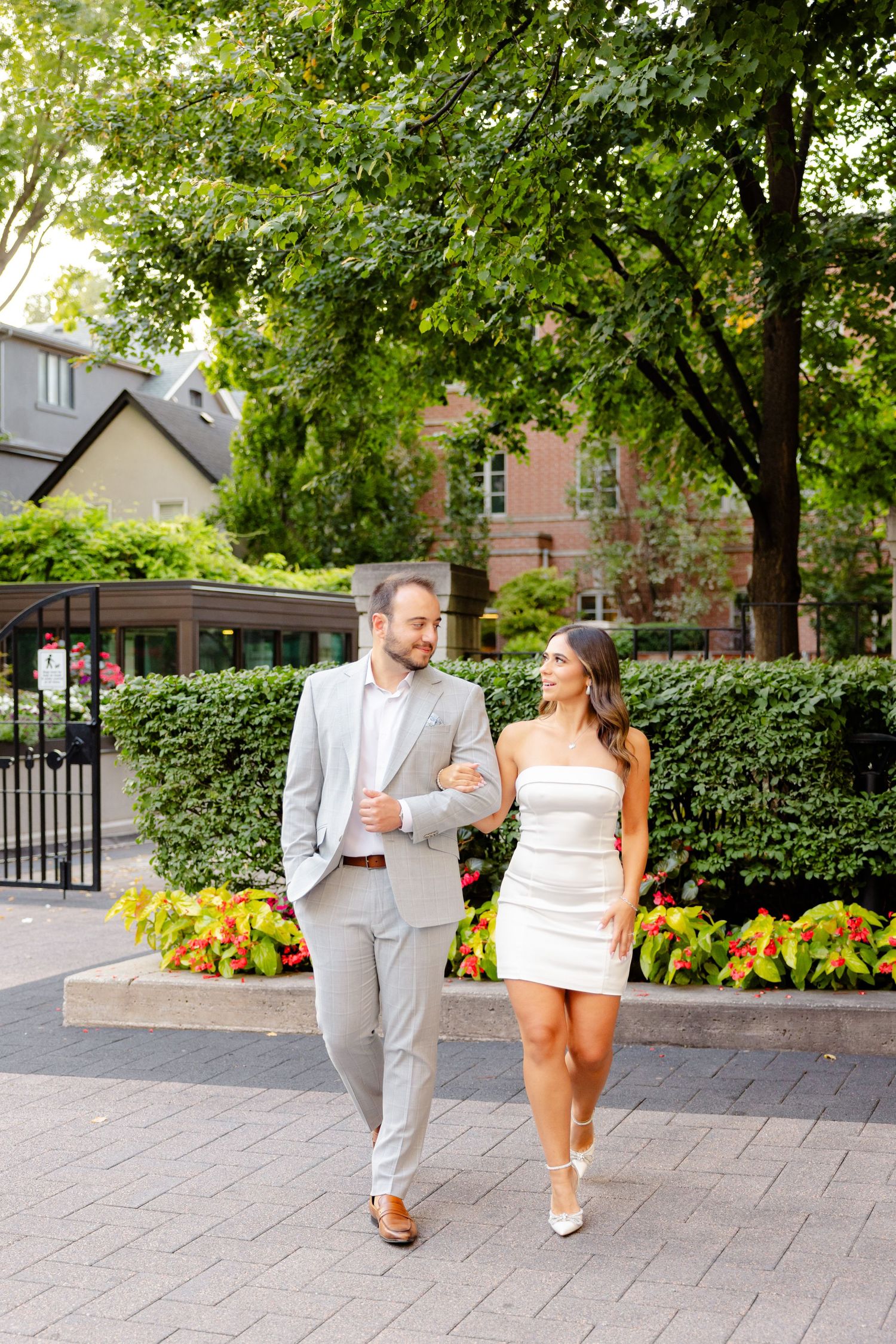 Couple in coordinated white and grey outfits stroll along a landscaped brick pathway on a sunny day.