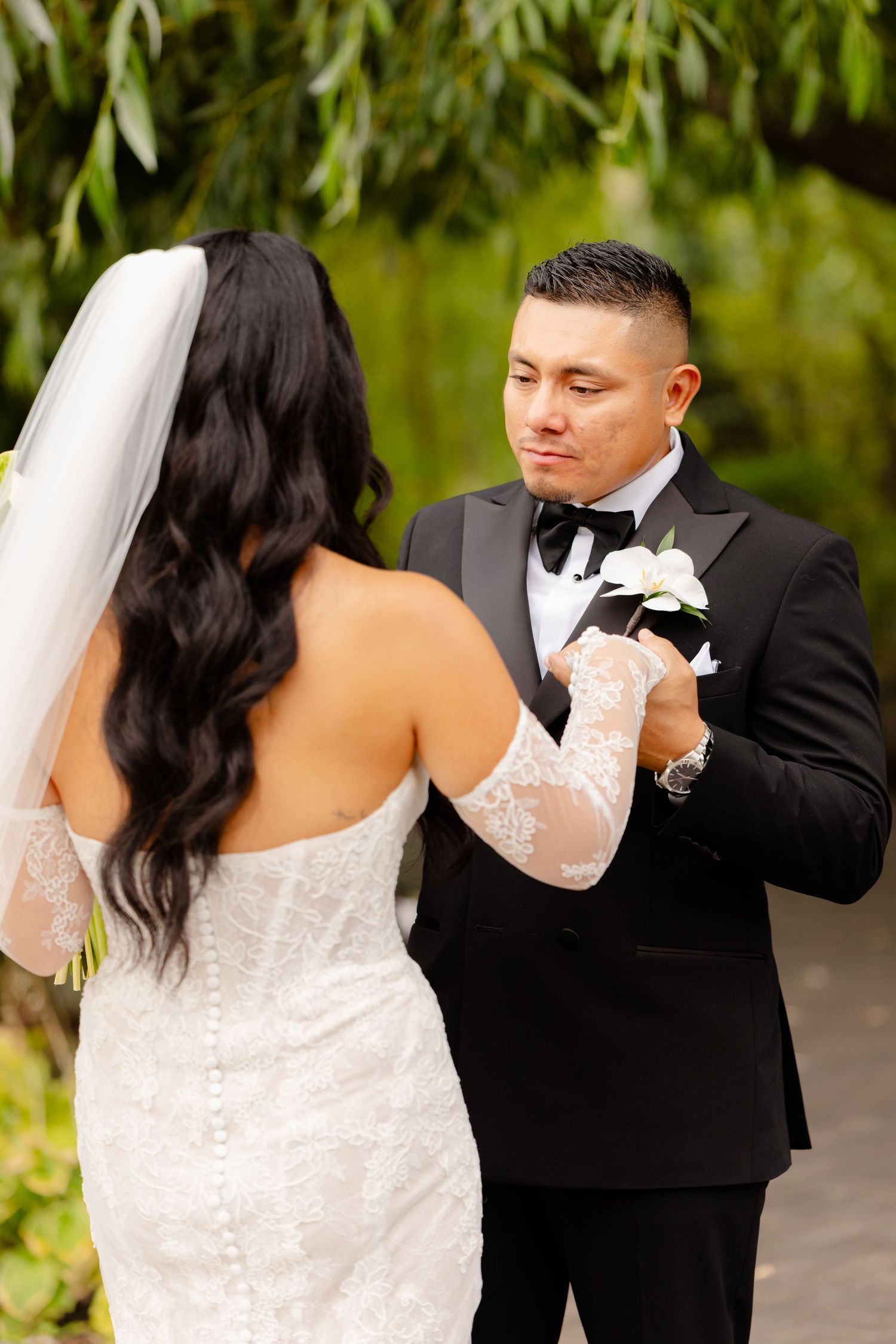 Couple in formal wedding attire and lace sleeves hold hands during outdoor ceremony with green foliage in background.