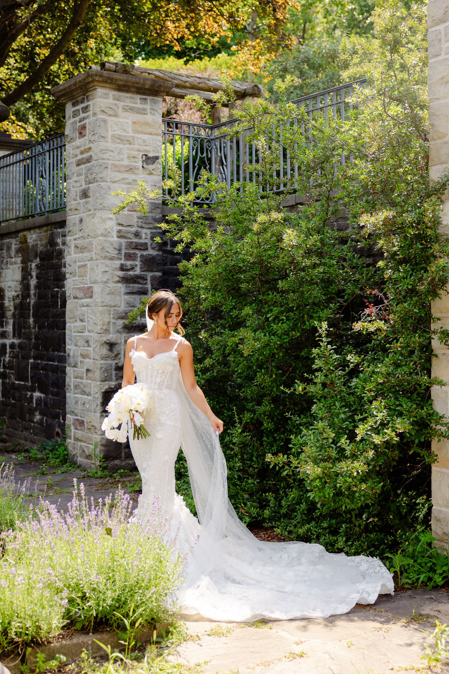 A bride in an elegant white mermaid gown with long train poses in a garden setting with stone walls and lavender blooms.