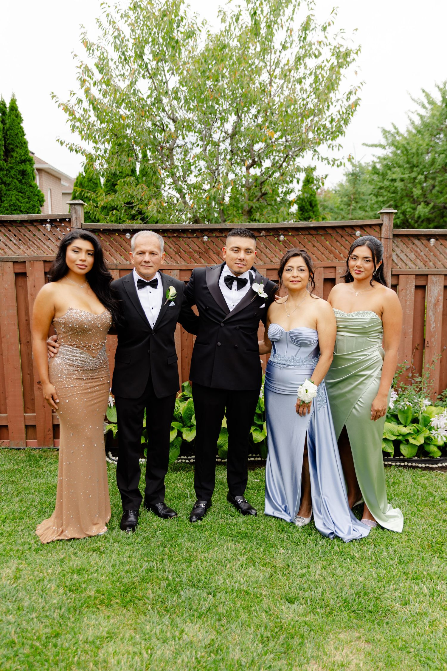 Wedding party members in formal attire pose together while standing on a green lawn near a wooden fence.