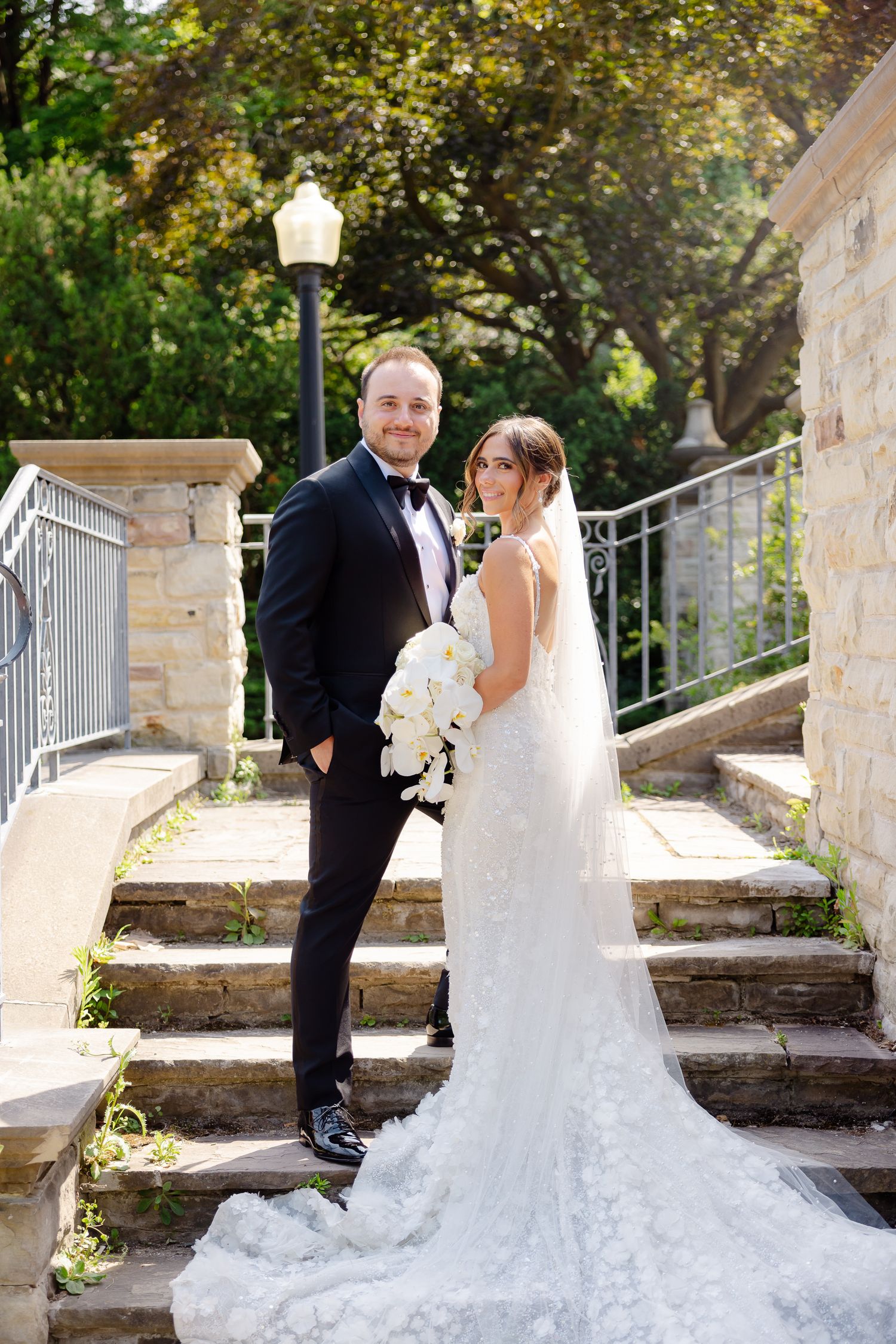 A couple poses on stone steps outdoors in elegant wedding attire with a long lace train and white bouquet.