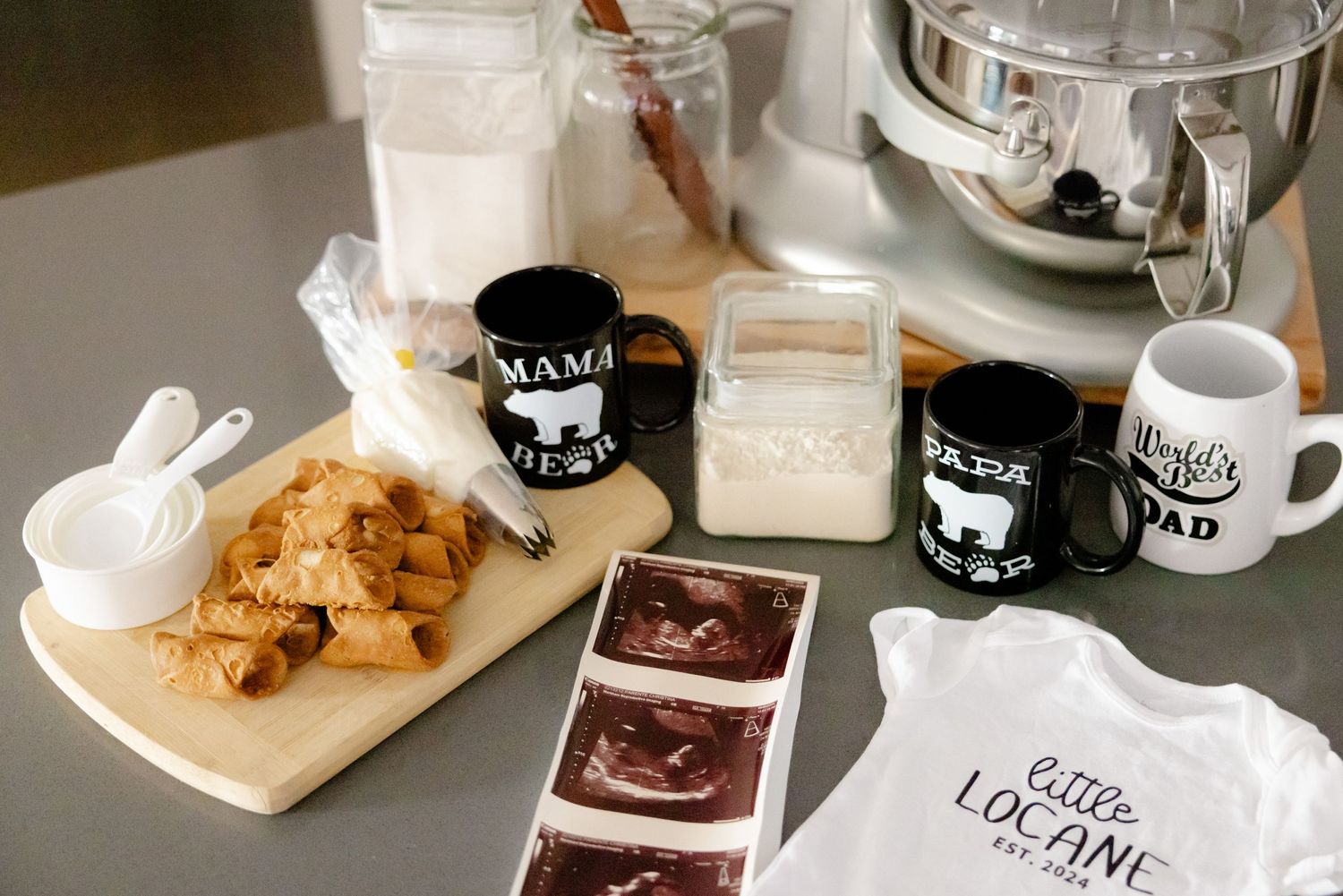 A table with mugs, cookies, milk, and baby-related items including ultrasound images and a onesie.