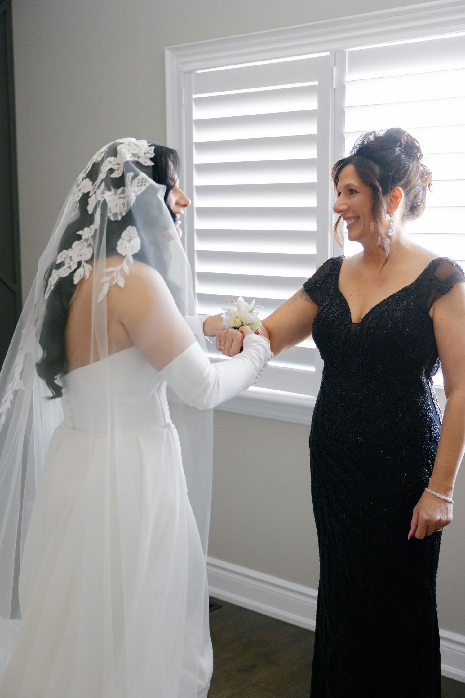 A bride in long white dress and veil holds hands with a woman wearing a black evening gown near a window.